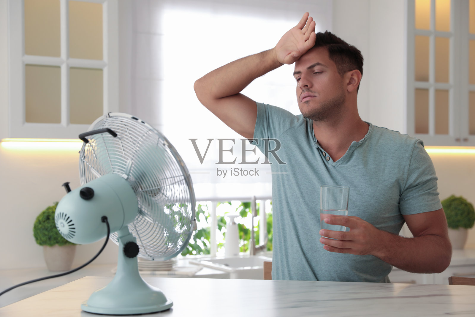 厨房的餐桌上有个男人,手里拿着一杯水和一把时髦的扇子。夏天热照片摄影图片