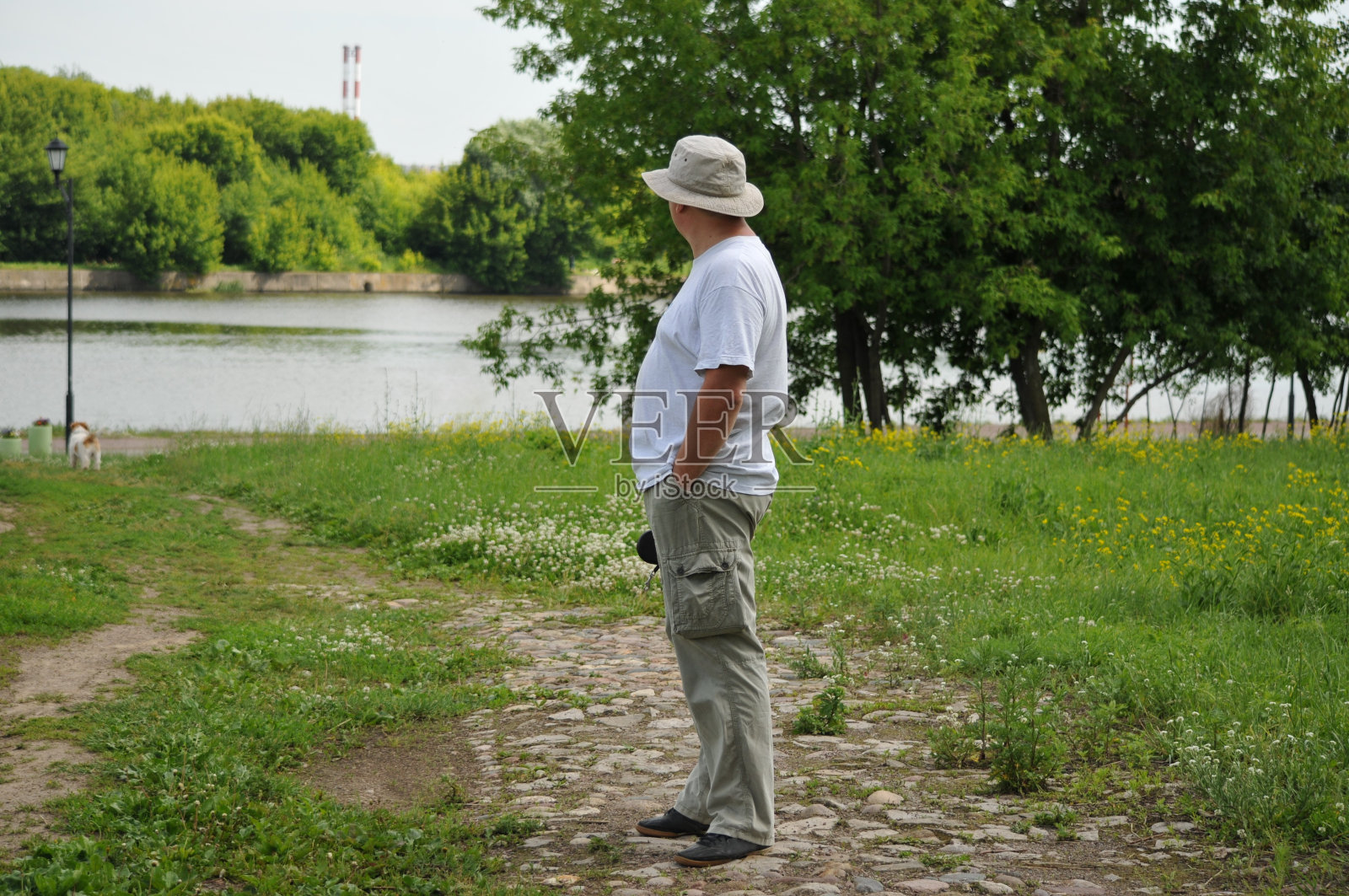 一个穿着白色巴拿马t恤和米色裤子的成年男子站在路上,手里拿着一条狗绳,看着河岸。照片摄影图片