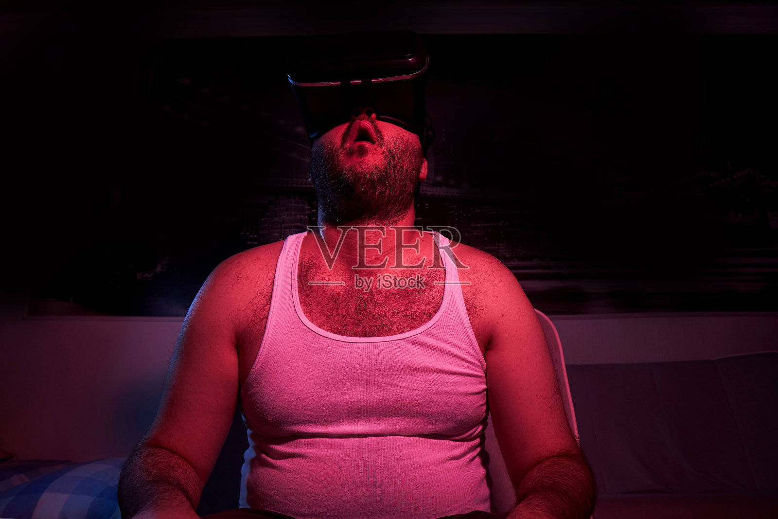 一名留着胡子的超重男子戴着VR眼镜看电视,表现出各种各样的情绪,他感到很惊讶。懒惰的中年男人照片摄影图片