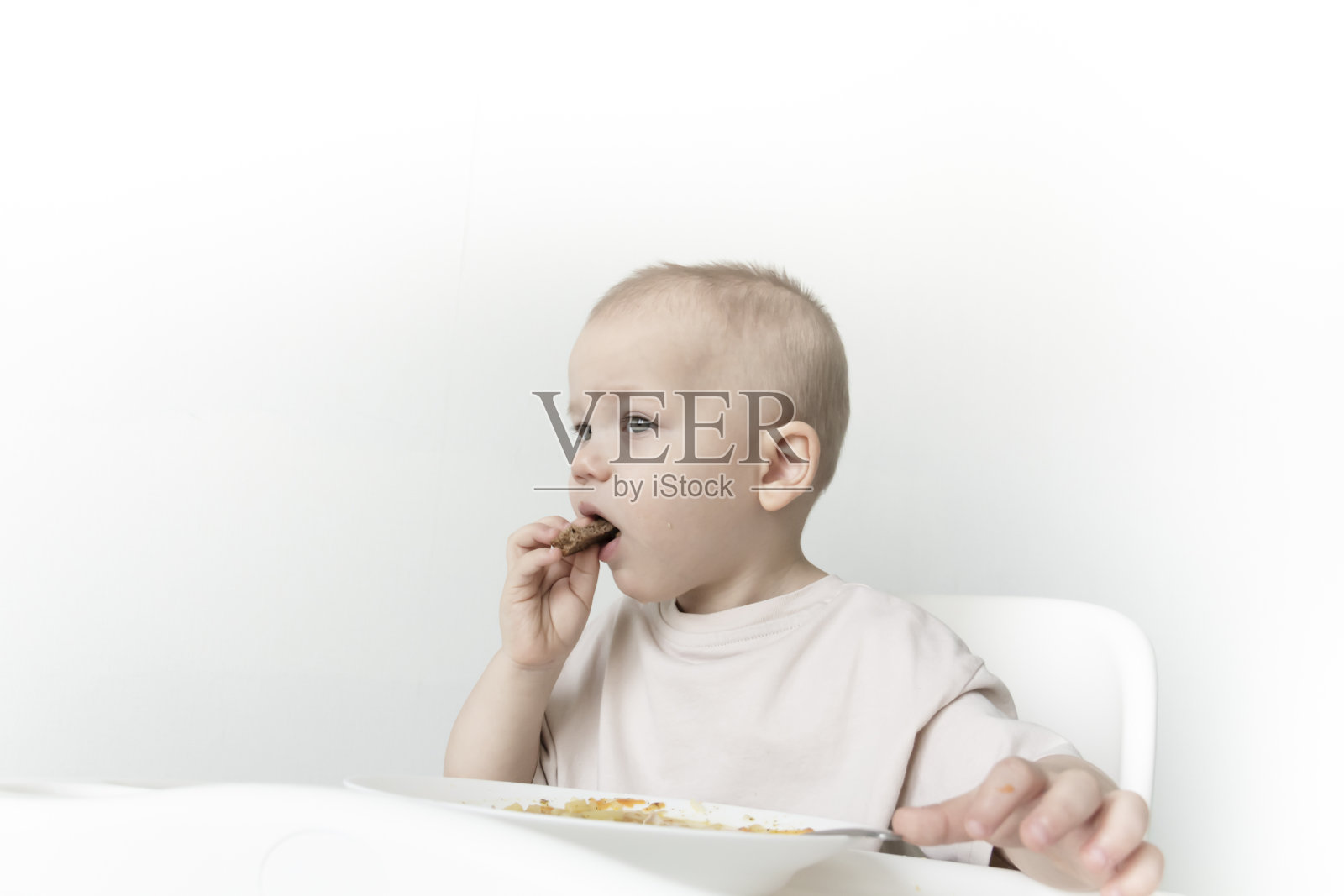 一个小男孩在白墙的背景下,坐在高脚椅上吃着面包和汤。照片摄影图片