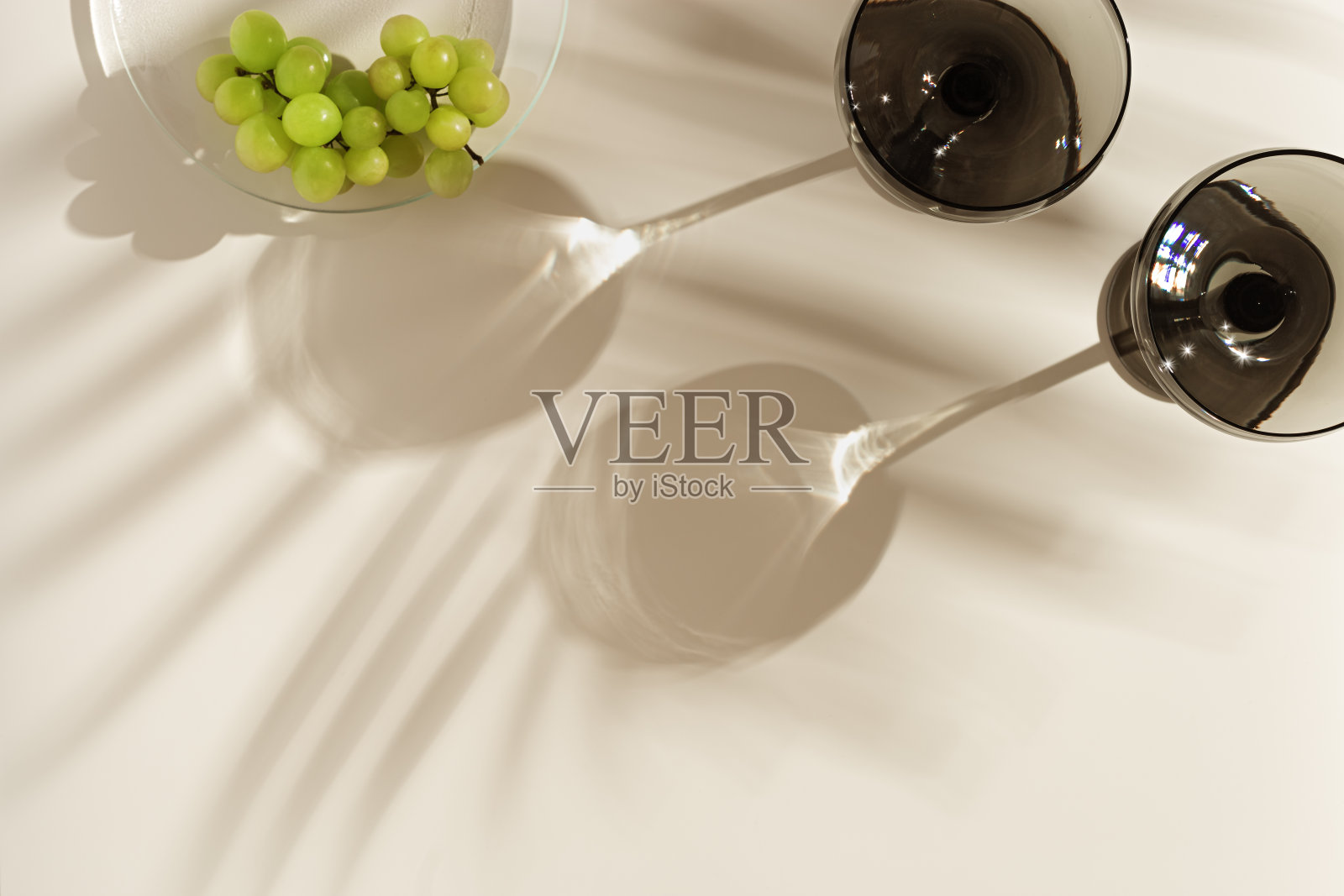 香槟玻璃着色灰色玻璃和葡萄米色背景与棕榈叶阴影,从太阳眩光。最小的夏季休息概念。用彩色玻璃器皿装的汽酒。创造性的俯视图照片摄影图片