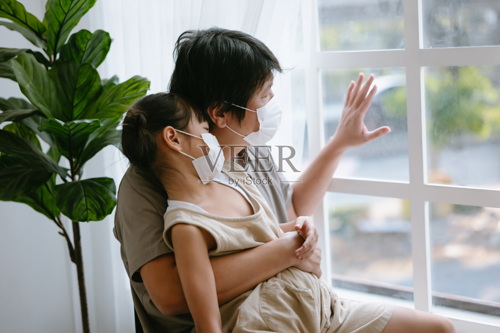 小亚洲女孩和她的父亲戴着医用口罩坐在椅子上,透过窗户看世界。悲伤的家人独自在家。封锁,COVID-19。冠状病毒爆发的情况。照片摄影图片