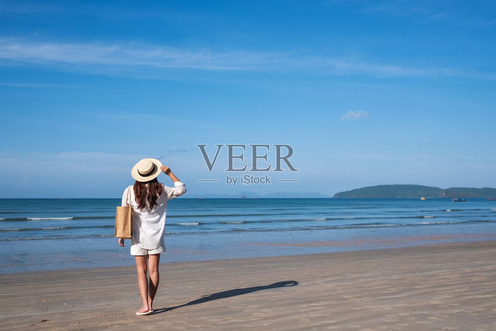 后视镜图像的一个年轻的女人,帽子和包走在蓝色的天空背景的海滩上照片摄影图片