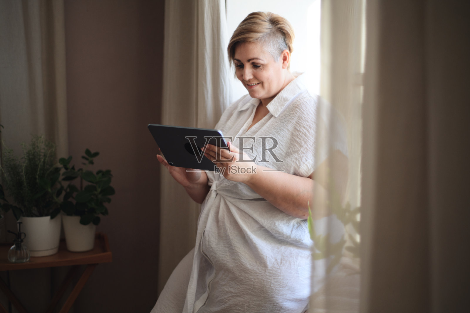 一个穿内衣的胖女人站在窗前用平板电脑。照片摄影图片