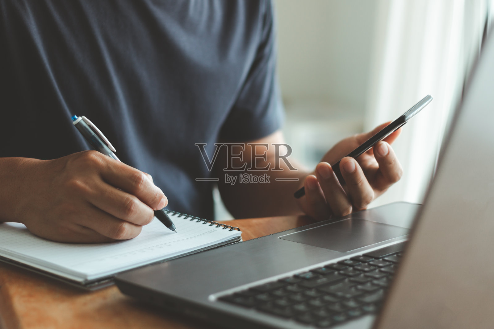 穿着t恤的商人坐在桌子旁,一边在客厅里工作,一边用智能手机在笔记本上写下信息。在家工作的概念。照片摄影图片