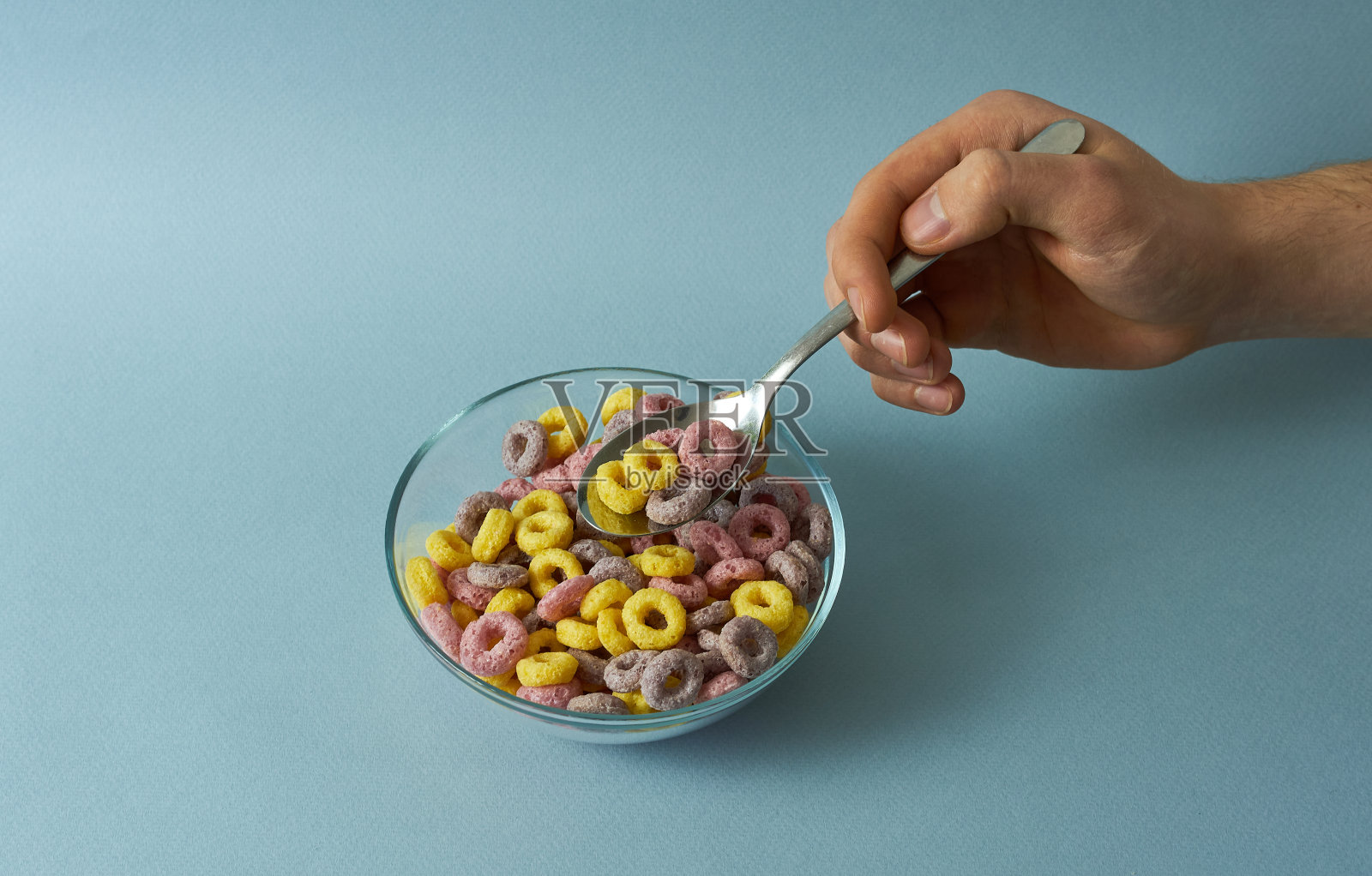 在蓝色的背景下,一个男人的手拿着一碗早餐麦片,勺子上有五颜六色的麦片圈照片摄影图片