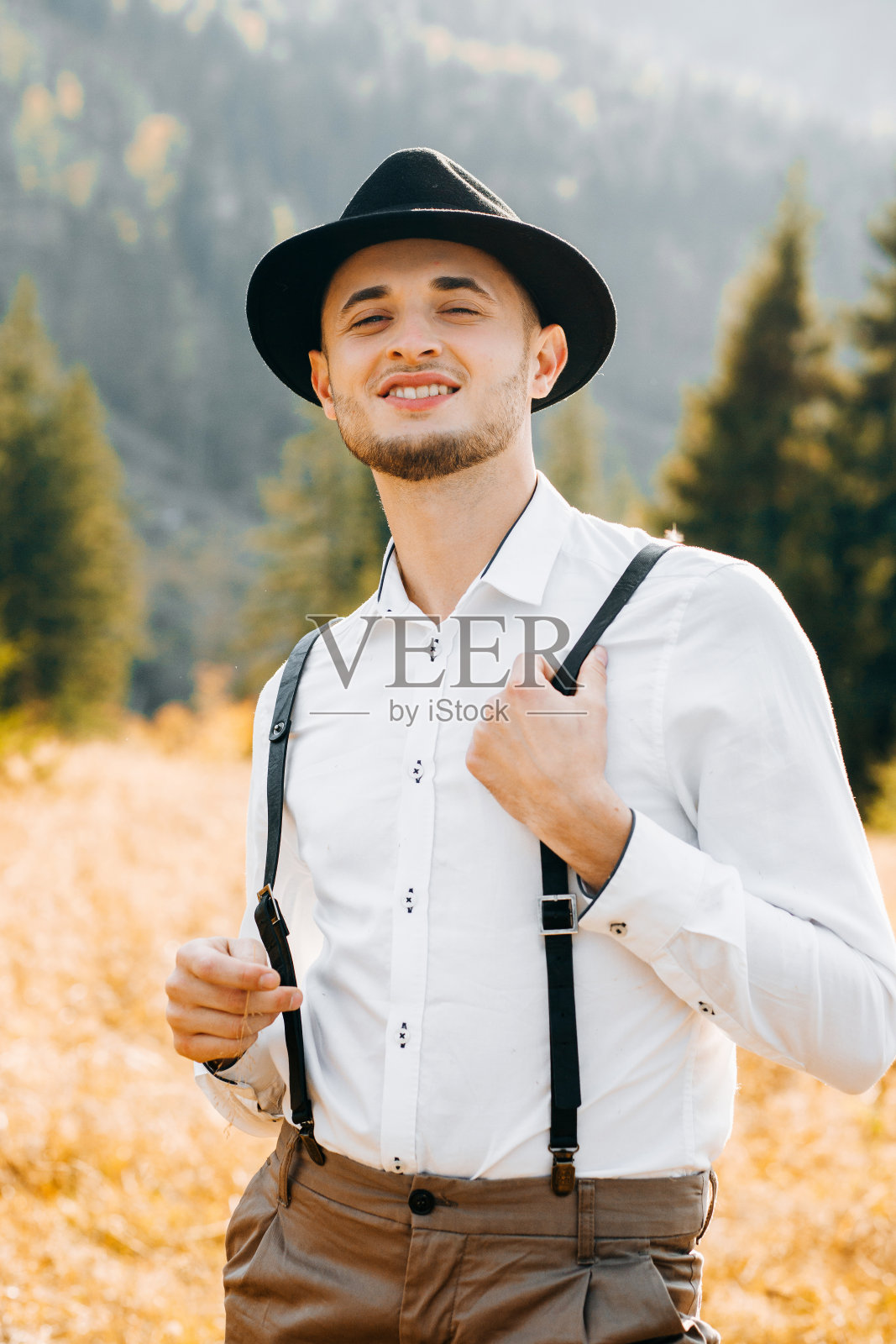 惊艳的新郎穿着白色衬衫,带着背带,戴着帽子,在山的背景上摆姿势。乡村风格照片摄影图片
