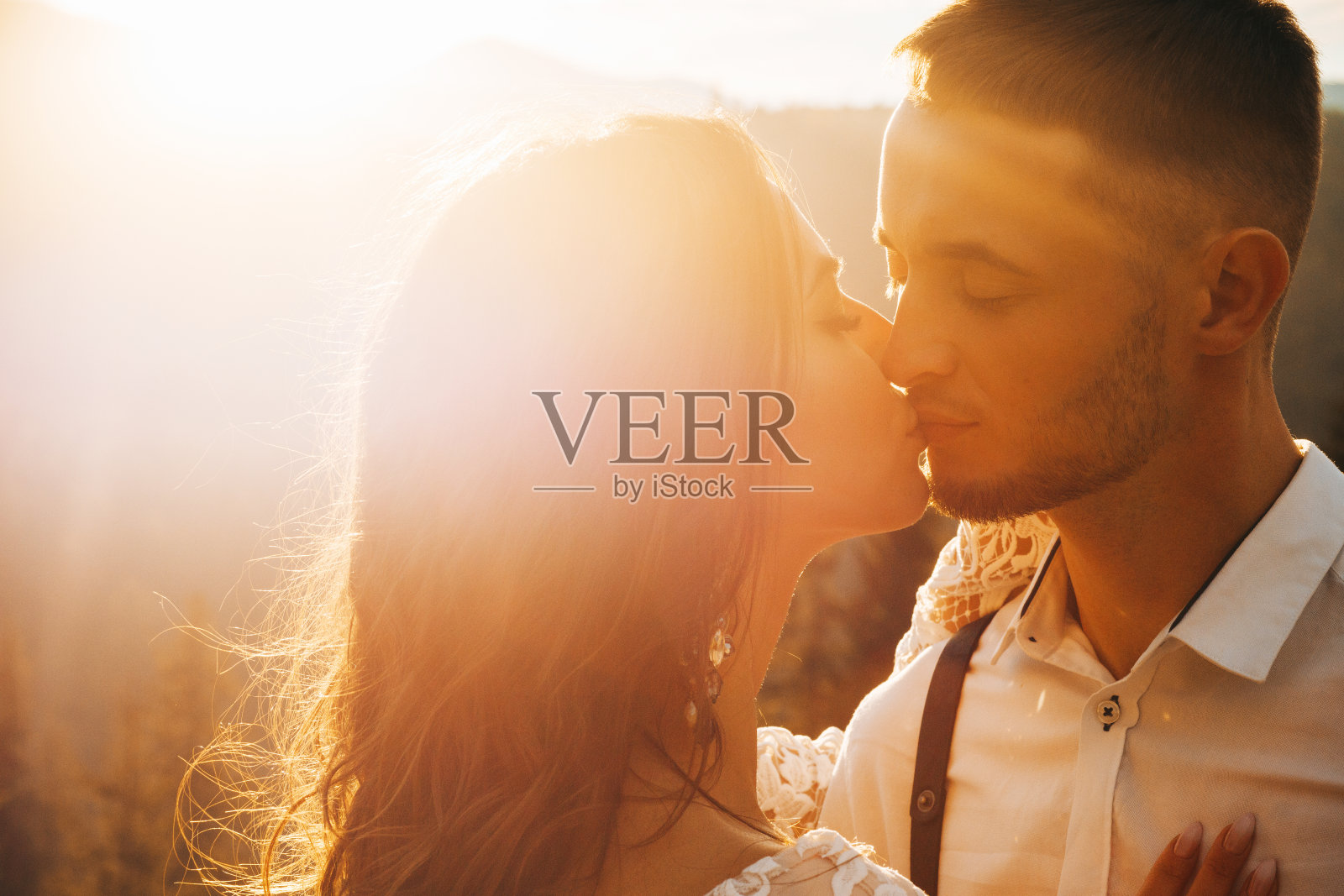 可爱的新郎和新娘在日落时分的山间摆着波西米亚式的姿势亲吻照片摄影图片