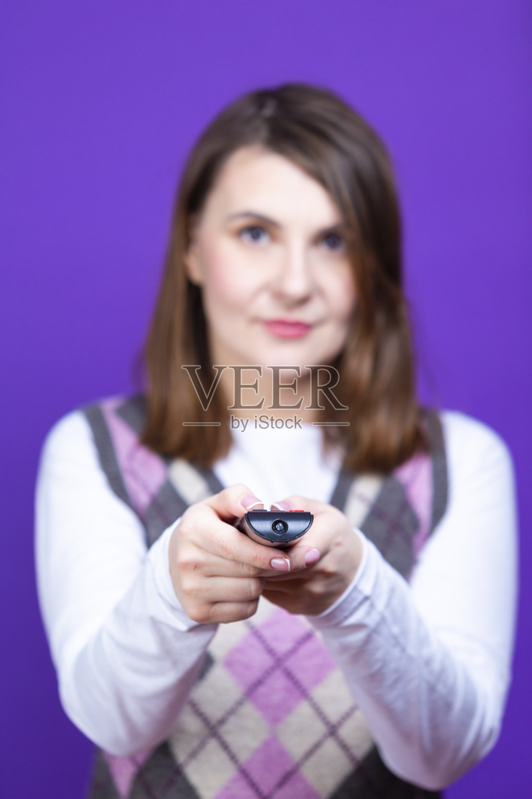 穿着紫色套衫的女性,拿着和指着电视遥控器,在紫色背景下摆姿势时,直接向前看照片摄影图片