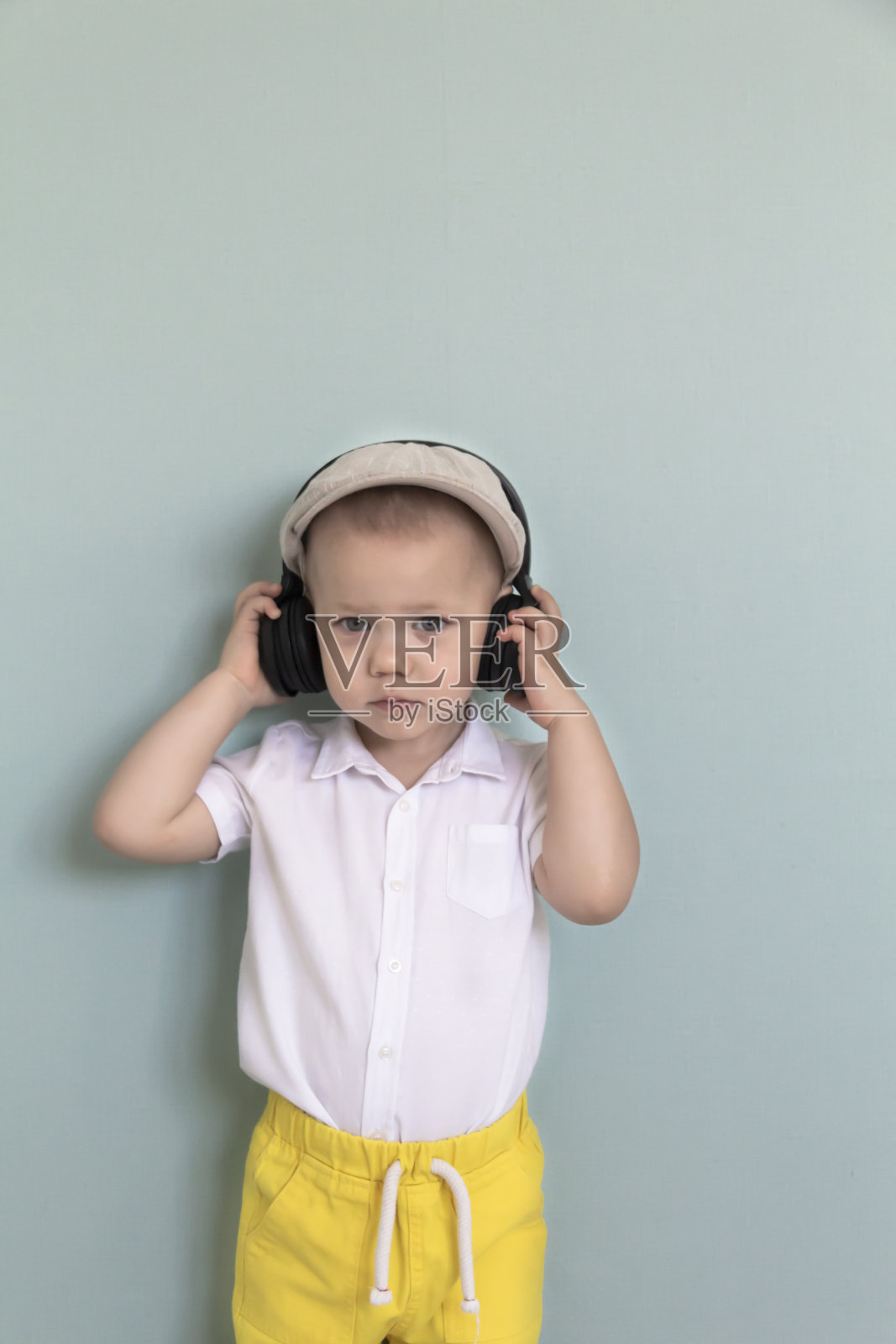一个小男孩穿着白色衬衫,黄色短裤,戴着一顶复古的帽子,戴着大耳机听音乐。照片摄影图片