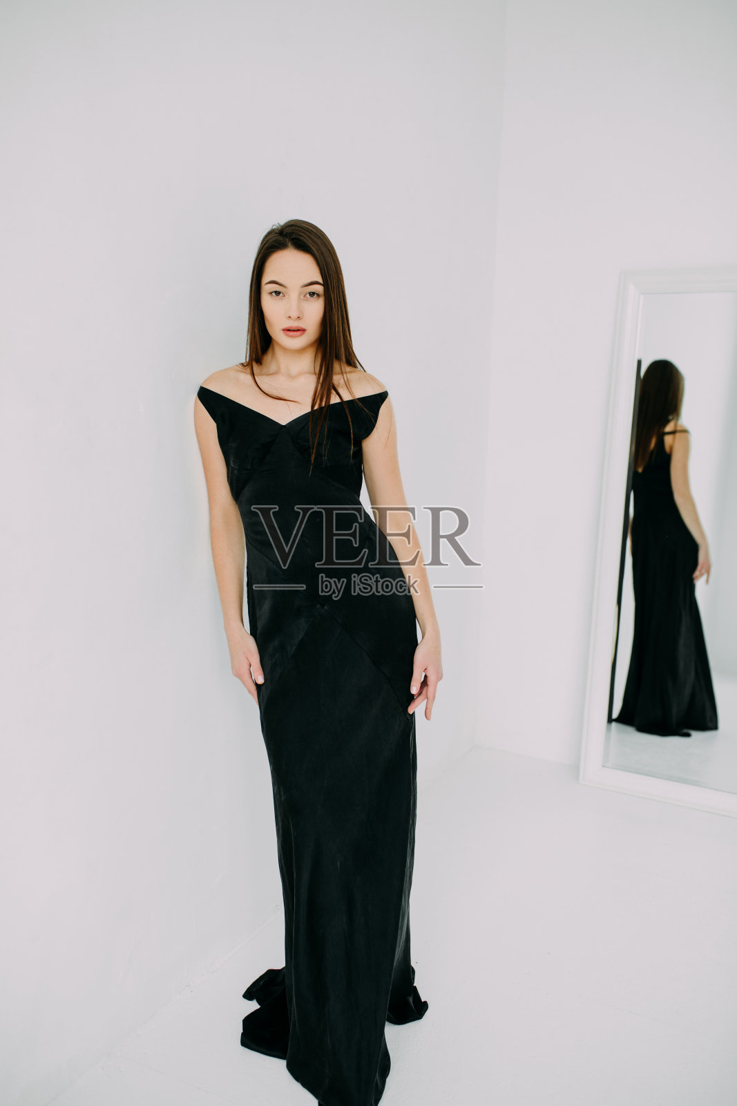 年轻苗条的黑发女人,穿着黑色长裙,站在镜子里她的倒影。照片摄影图片
