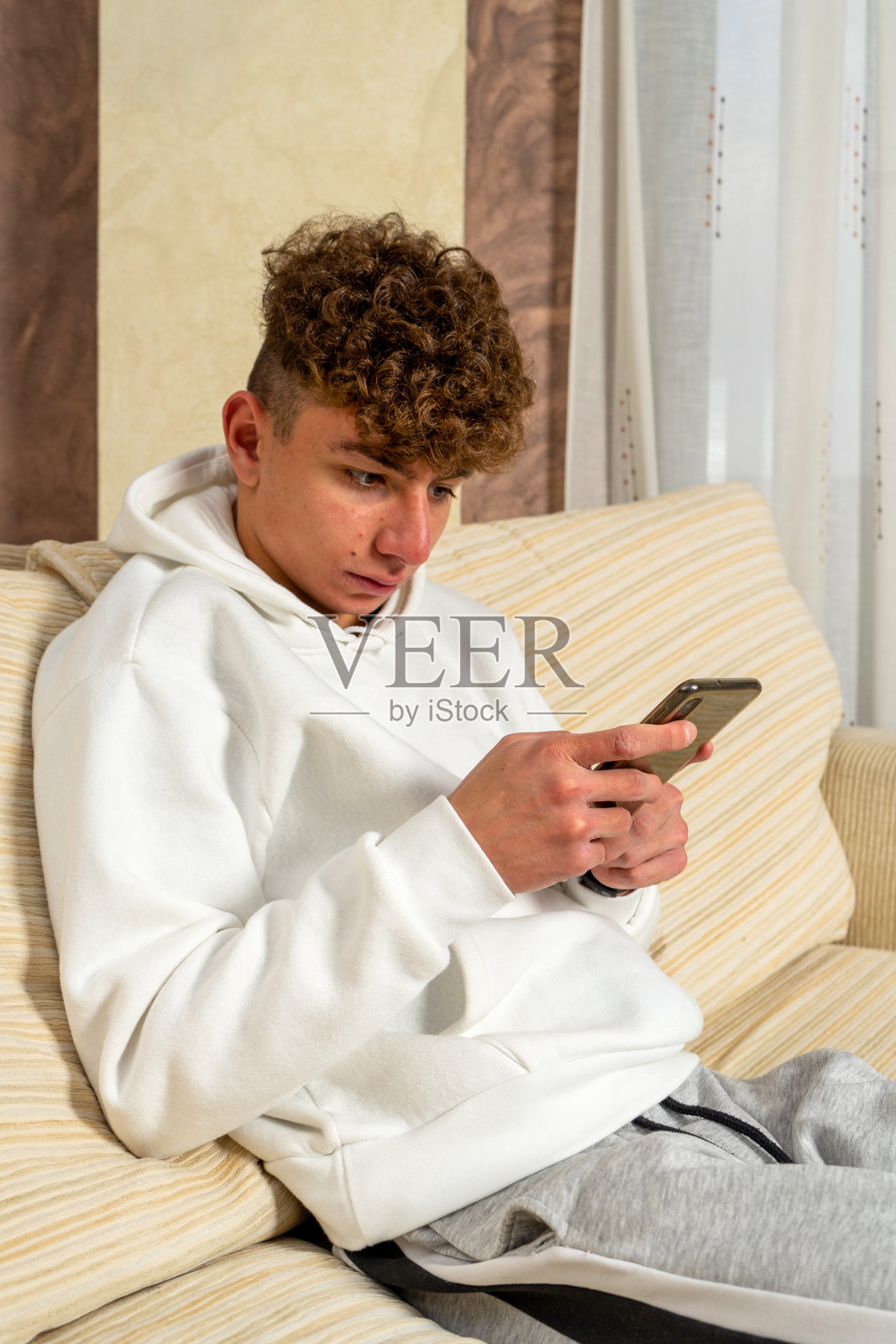 一头卷发、穿着运动服的白人年轻人坐在白色沙发上,用智能手机打字,给社交网络上的朋友们写信照片摄影图片