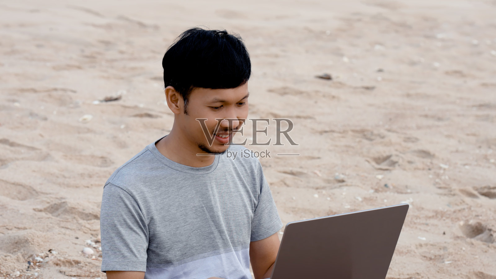 在海边,凉爽的海浪拍打着他,英俊的年轻亚洲男子在他的笔记本电脑上与主管讨论工作。照片摄影图片