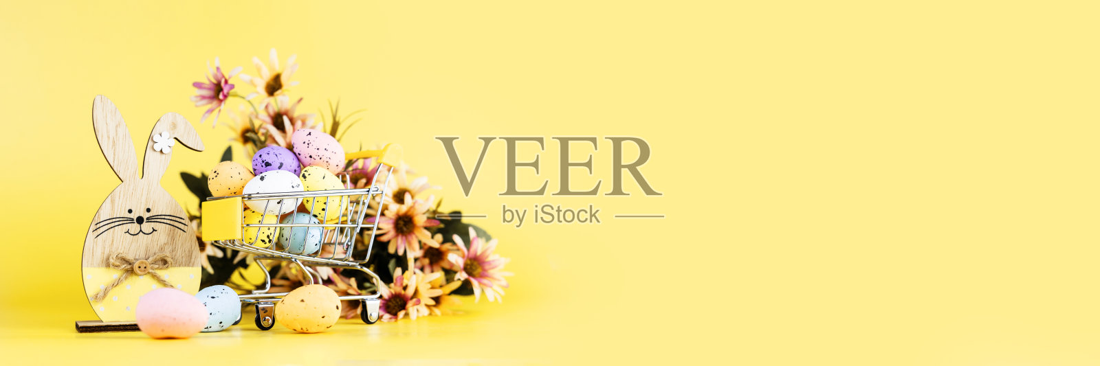 五颜六色的复活节鹌鹑蛋在购物车,木制兔子和春天菊花在黄色的背景。副本的空间。最小的复活节的概念。横幅照片摄影图片