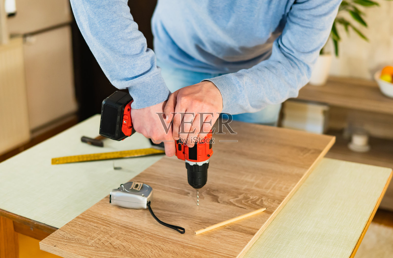 一个年轻人拿着钻头在他家里桌子上的木板(胶合板)上钻洞的特写镜头。在家DIY项目。照片摄影图片