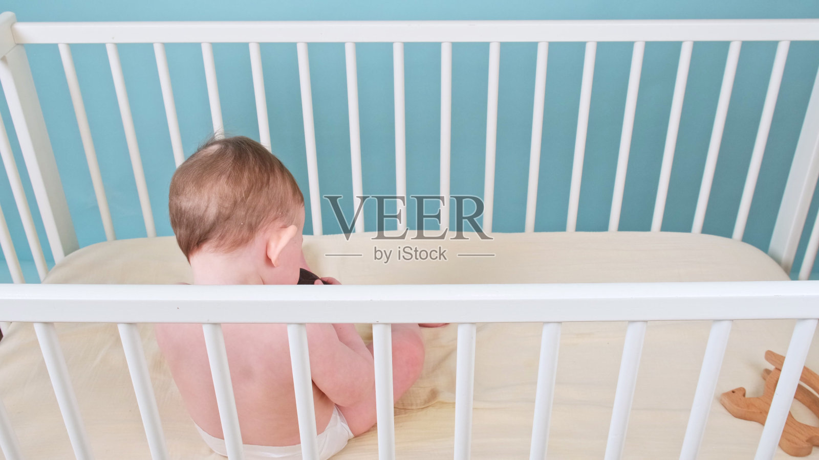 小男孩坐在婴儿床上玩玩具,蓝色的画室背景。穿着白色尿布的白人小孩照片摄影图片