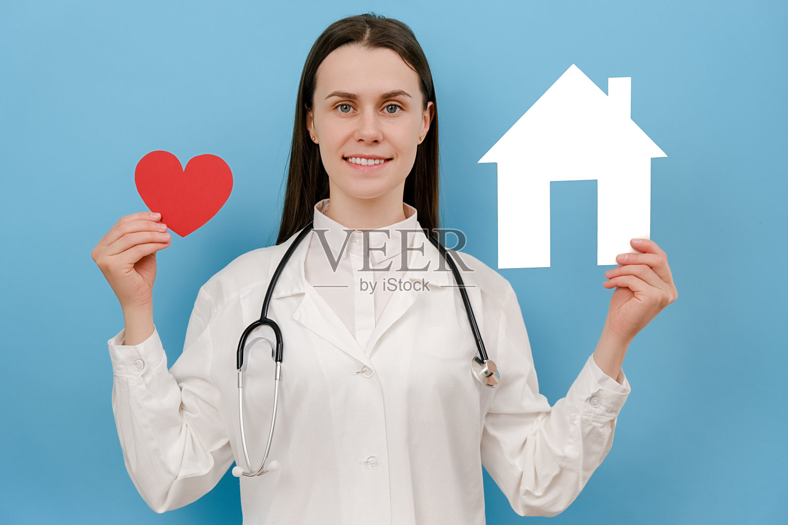 快乐的年轻女医生穿着专业的医疗白色制服和听诊器抱着小房子和红心,看着摄像机,孤立在蓝色的工作室背景上。关怀、药物、帮助和支持照片摄影图片