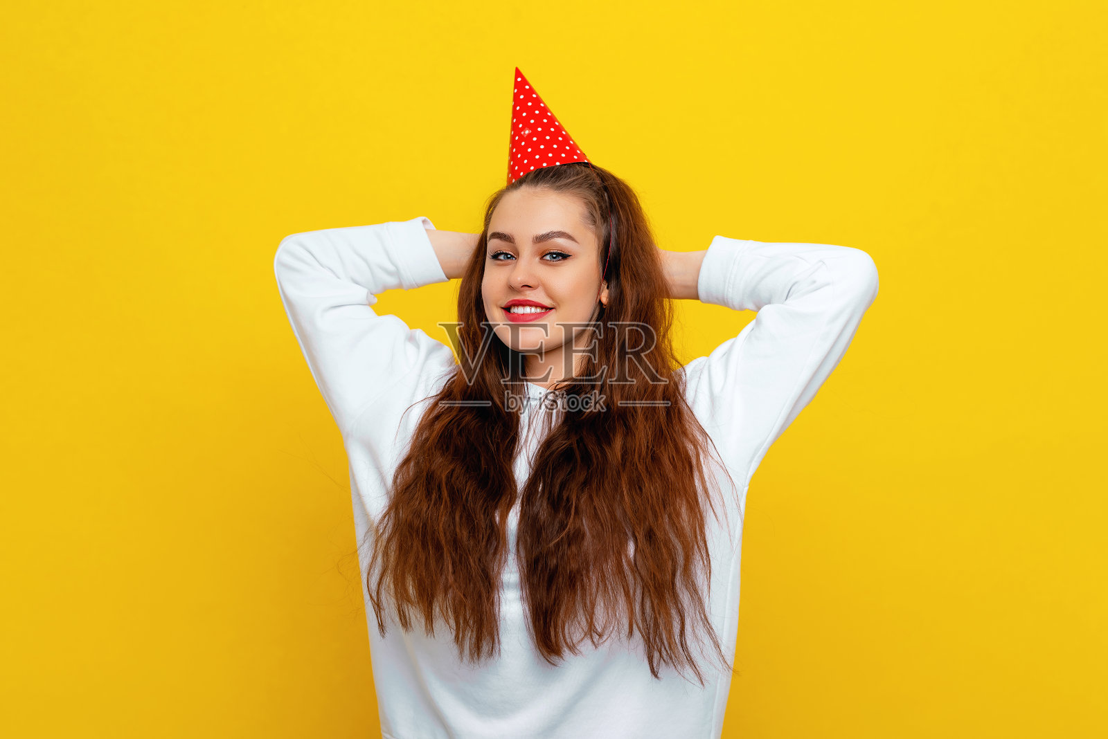 美丽有趣的年轻快乐的女人在白色休闲风格运动衫和生日派对帽子迷人的微笑和棕色头发摆姿势在黄色的背景照片摄影图片