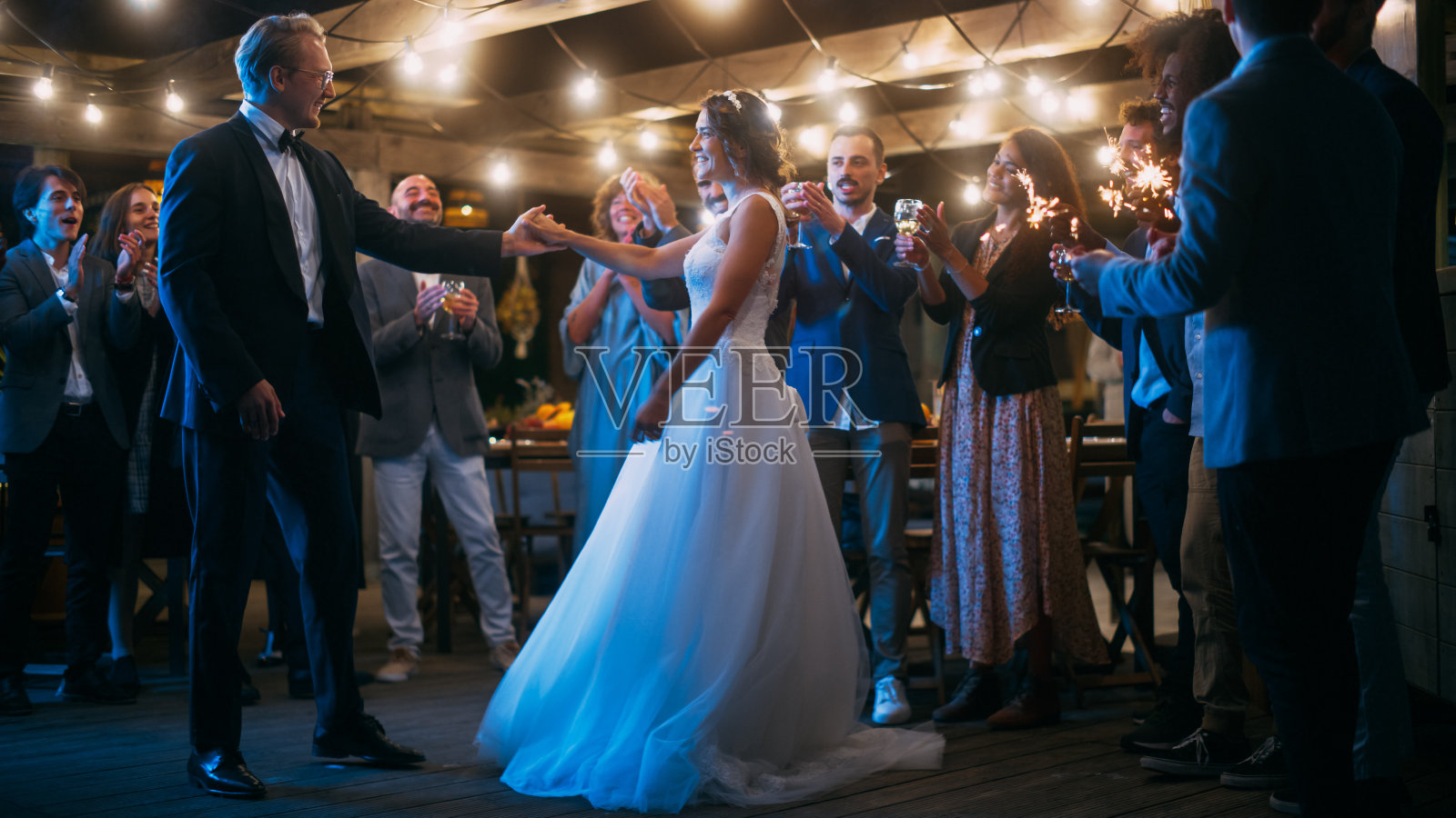 美丽的新娘在白色礼服和新郎在时尚的黑色西装庆祝婚礼在一个晚上的接待派对。新婚夫妇与来自不同民族的朋友在一起跳舞。照片摄影图片