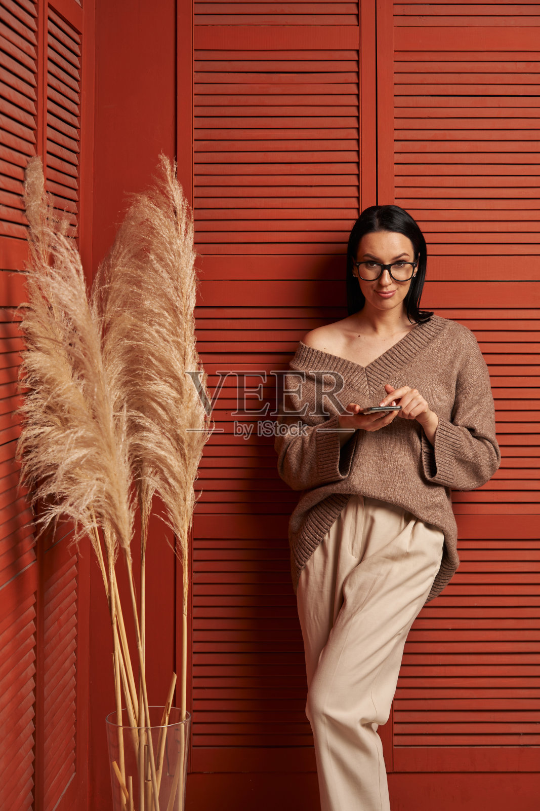 一位美丽的年轻女子,一头黑发,戴着眼镜,穿着舒适的衣服,在红色板条墙的背景下看着智能手机里的东西,旁边是花瓶里高高的潘pas草照片摄影图片