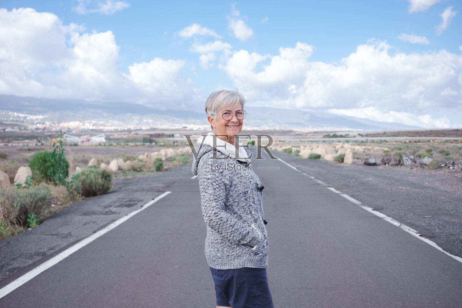 微笑独自旅行者年长妇女走在户外享受自由看着相机。空荡的道路上,苍茫的山峦和蔚蓝的天空上,有一位成熟的老妇人照片摄影图片