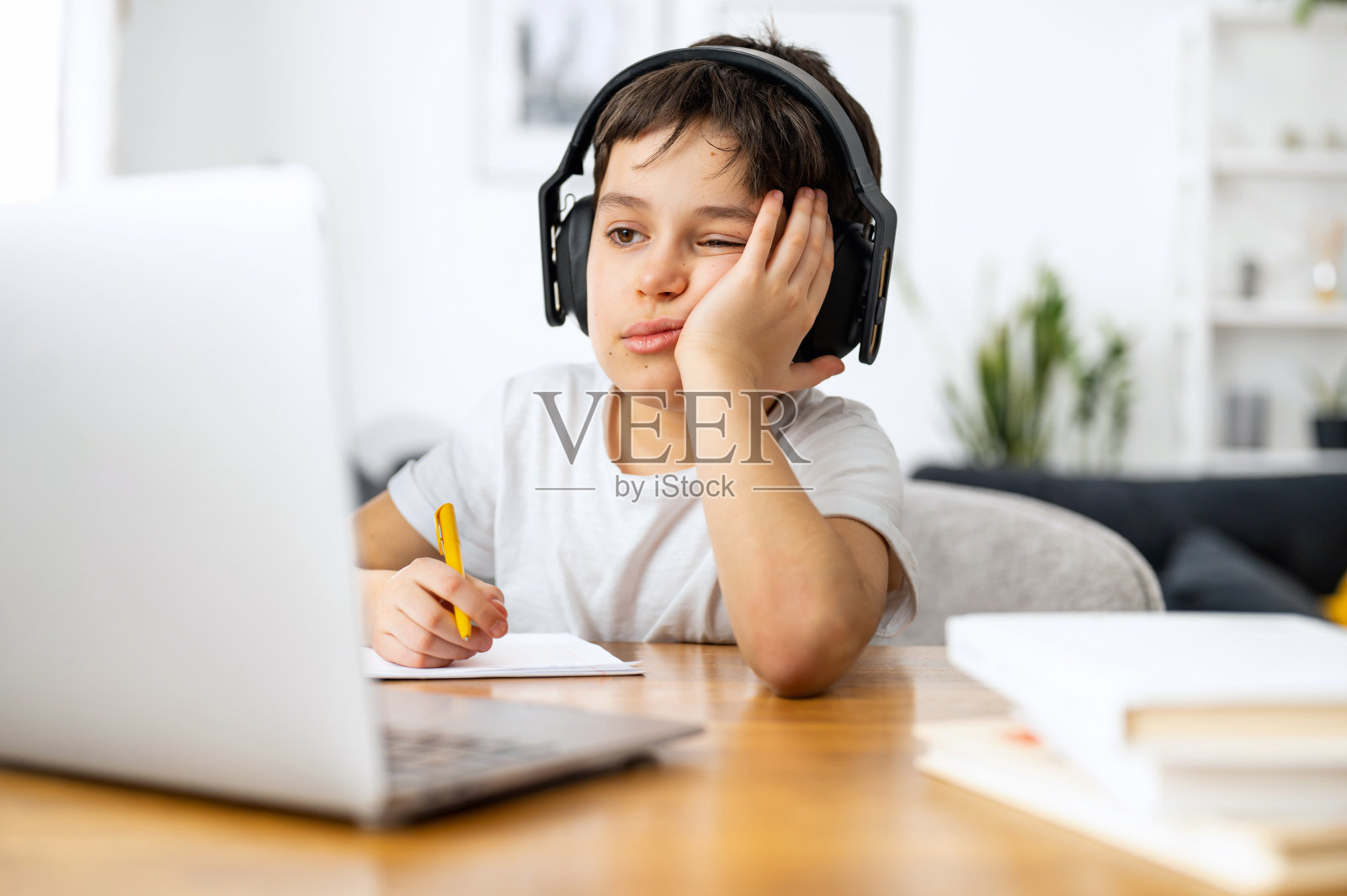 疲惫的学生做作业,戴着耳机听音乐,远离家学习,使用时髦的笔记本电脑,青春期前的男孩在笔记本上写任务照片摄影图片