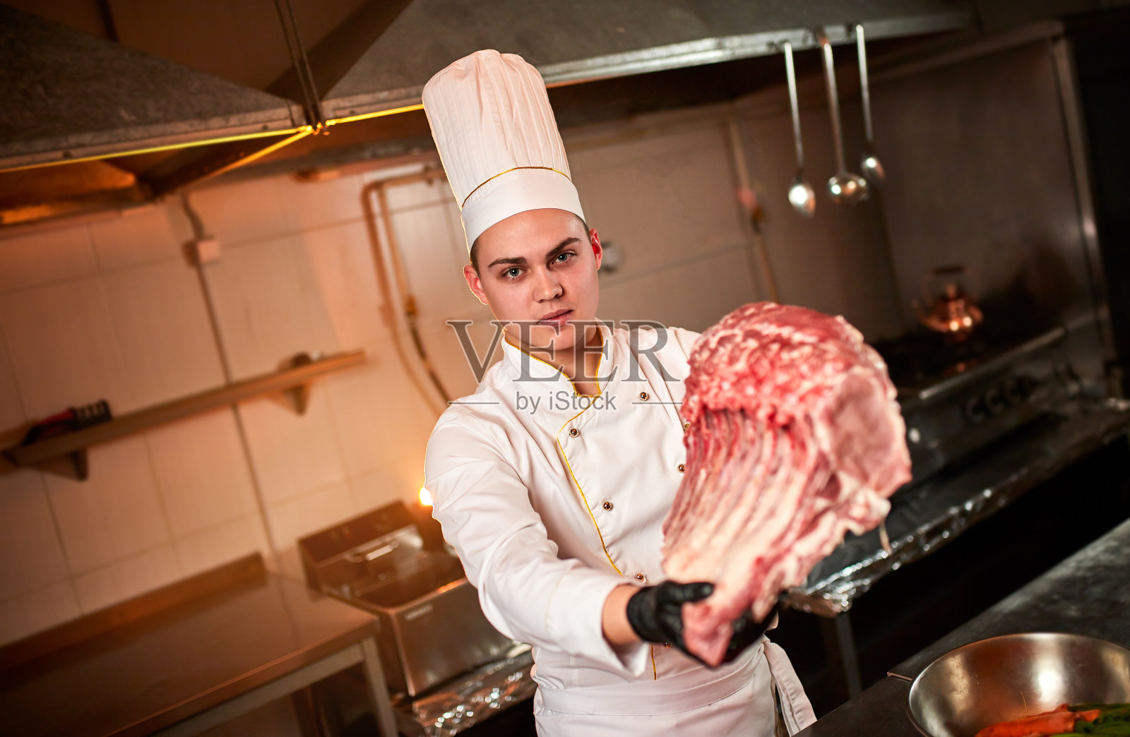 专业厨师拿着生的烤肉排骨放在蔬菜的背景上。厨师长在餐厅厨房准备烤肉照片摄影图片