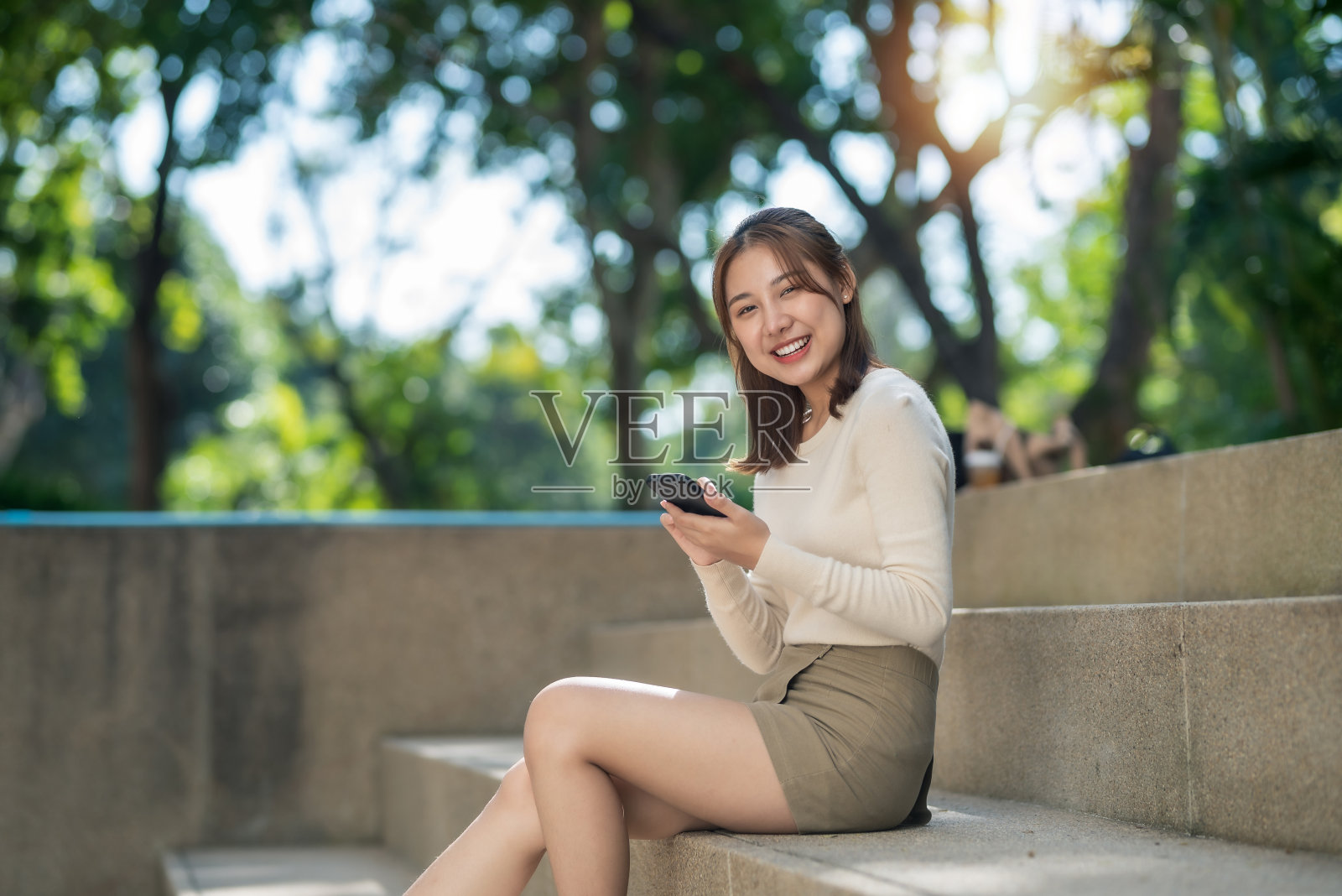 微笑快乐的年轻亚洲学生坐在外面的大学台阶上用手机。看相机。照片摄影图片