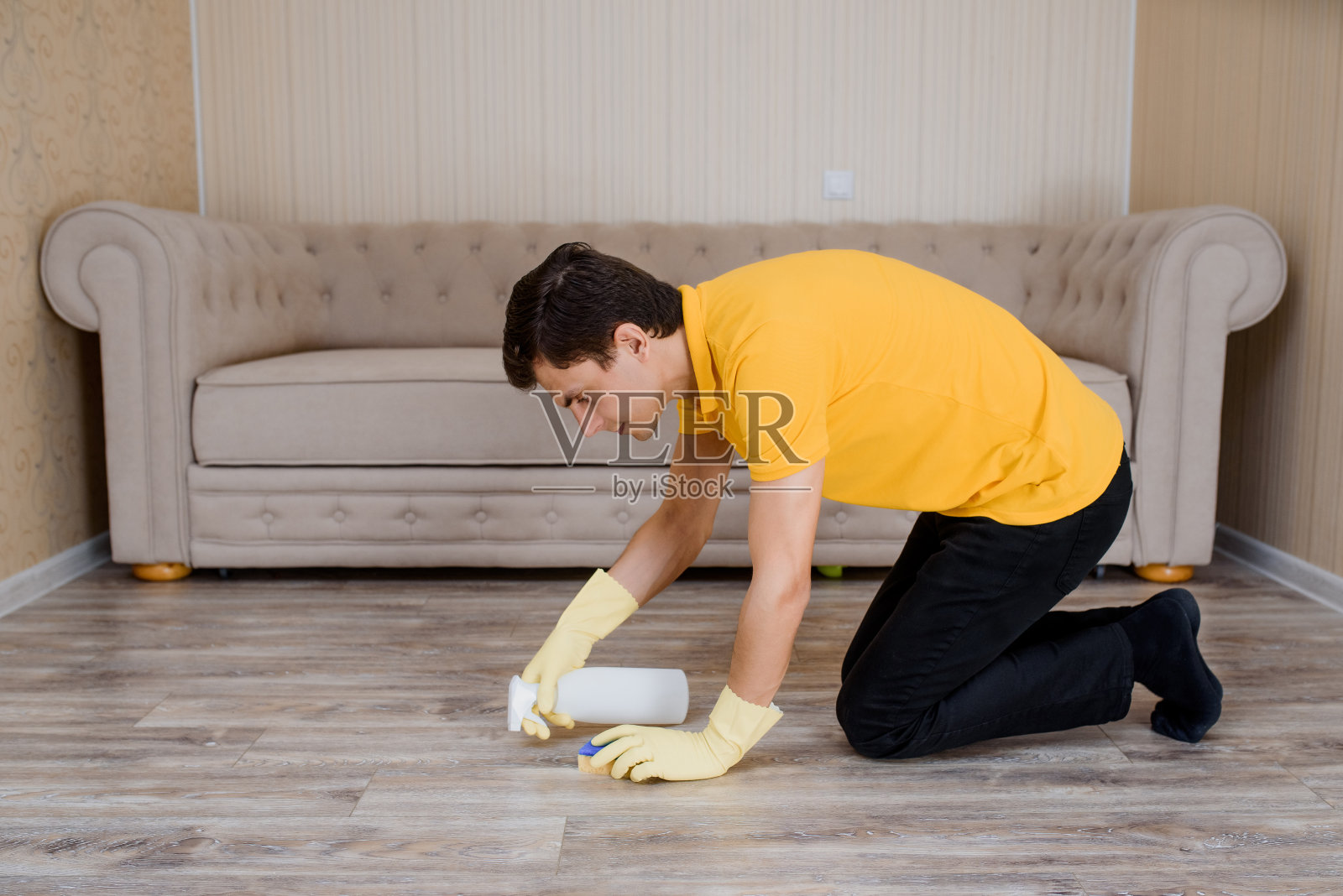 身穿黄色t恤、戴着黄色手套的男子正在用清洁喷雾和海绵清洁木地板。照片摄影图片