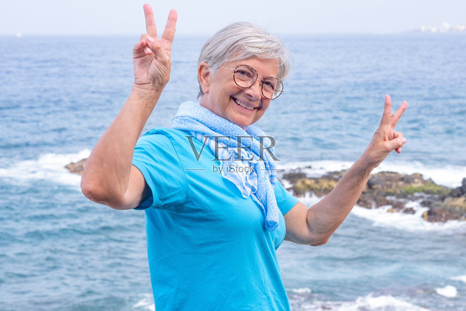 迷人的肖像快乐的年老妇女穿着蓝色t恤和围巾享受户外度假在海上。漂亮的女人看着镜头用手做出胜利的手势照片摄影图片