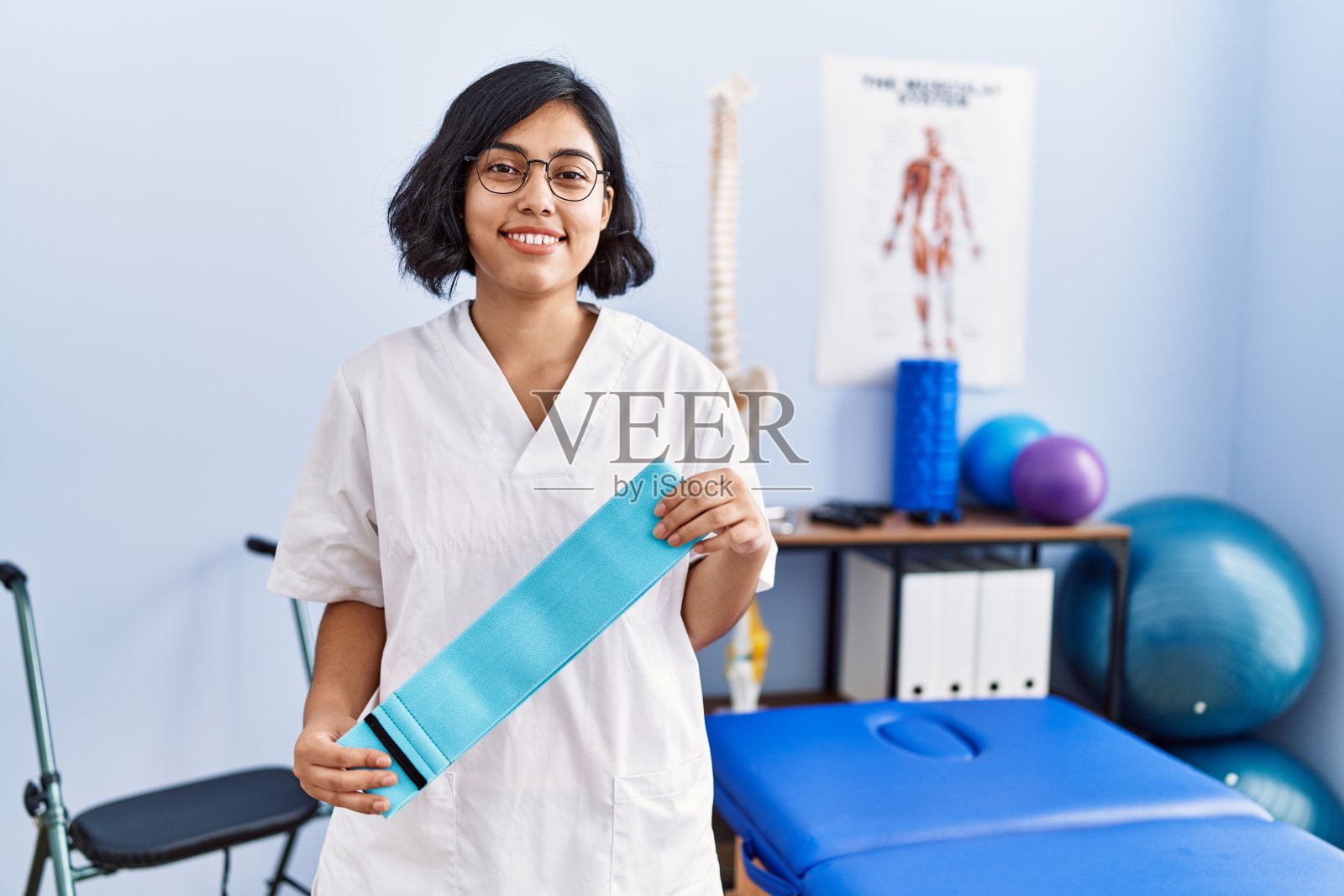 年轻的拉丁妇女穿着物理治疗医师制服持有橡皮筋在物理治疗诊所照片摄影图片