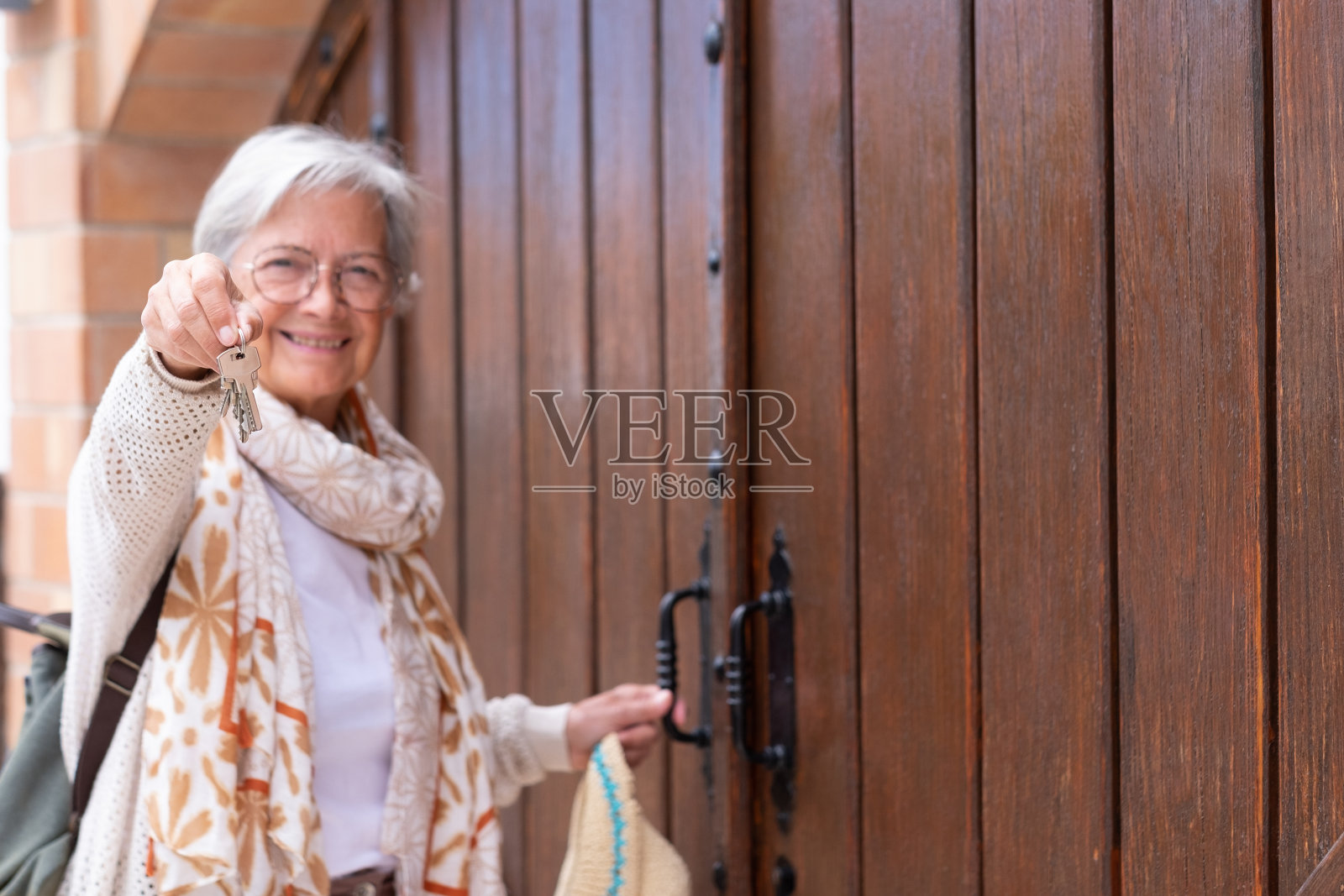 散焦微笑的老妇人看着相机打开一个旧的巨大的木屋门,同时持有家的钥匙照片摄影图片