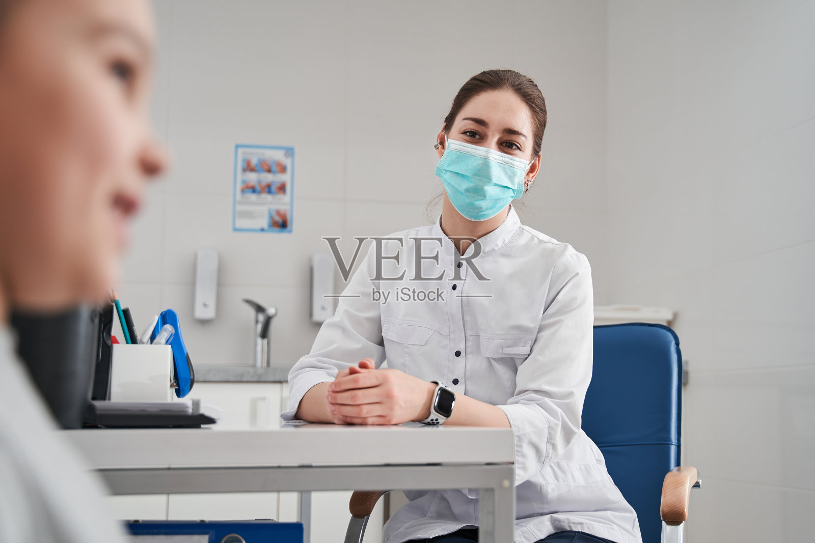 戴着防护口罩的医生坐在桌旁和她的小病人交谈照片摄影图片