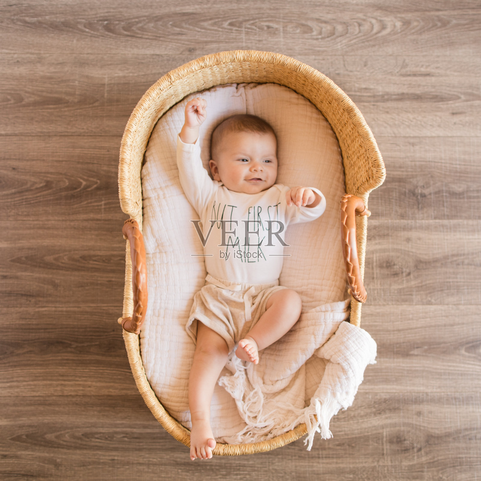 一个快乐的14周大的男婴穿着白色的连体衣,上面写着“但第一次牛奶”,躺在一个舒适的奶油棉毯子在海草摩西篮子里照片摄影图片