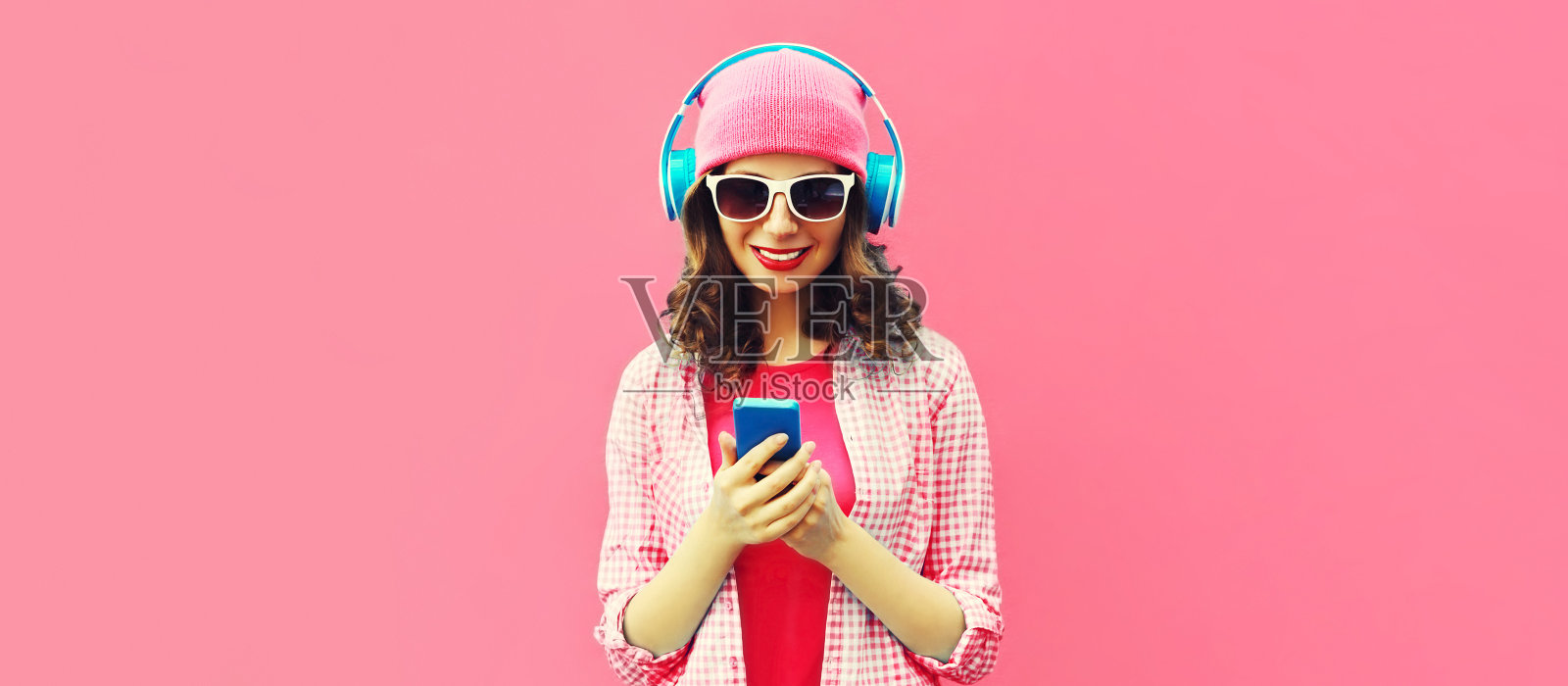 夏天五颜六色的肖像时尚的现代年轻女子听音乐耳机与智能手机在粉红色的背景照片摄影图片