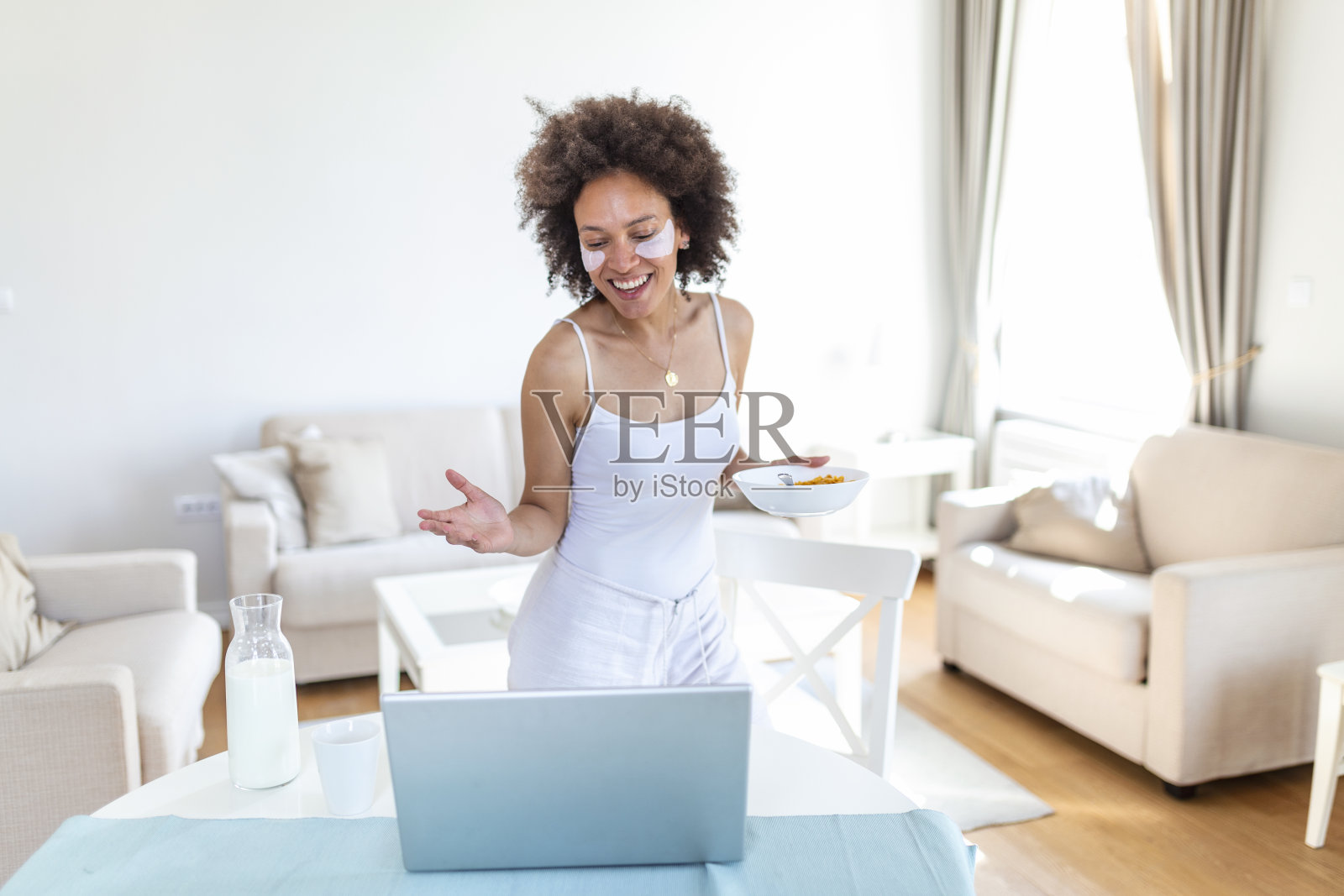 年轻迷人的女人坐在室内的桌子旁,手提电脑上放着玉米片。看着笔记本电脑和她的朋友通过视频通话。照片摄影图片