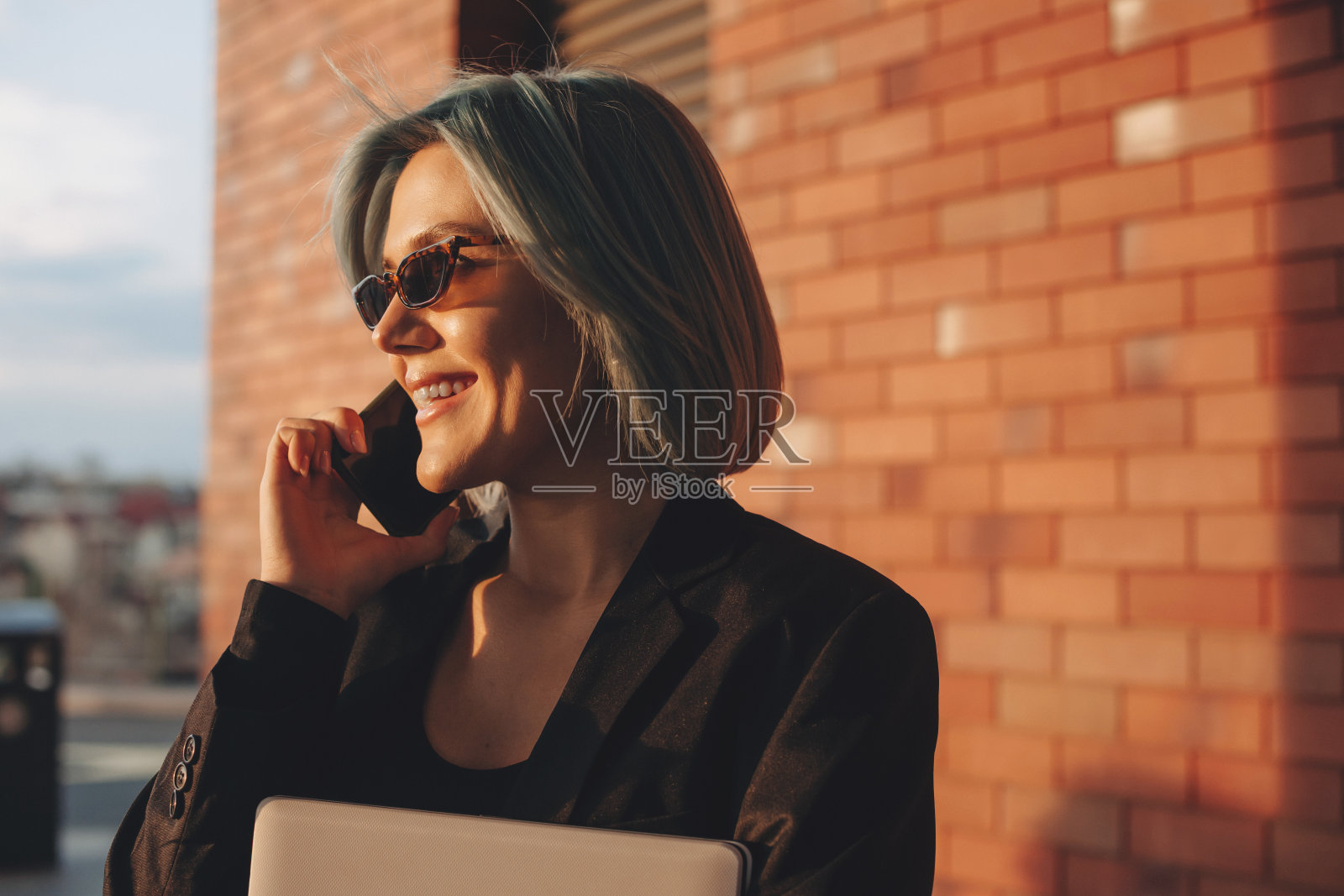 蓝色头发的女人站在外面的砖墙上,微笑着用智能手机聊天。移动电话。移动通信。说话的人。商业的女人。照片摄影图片