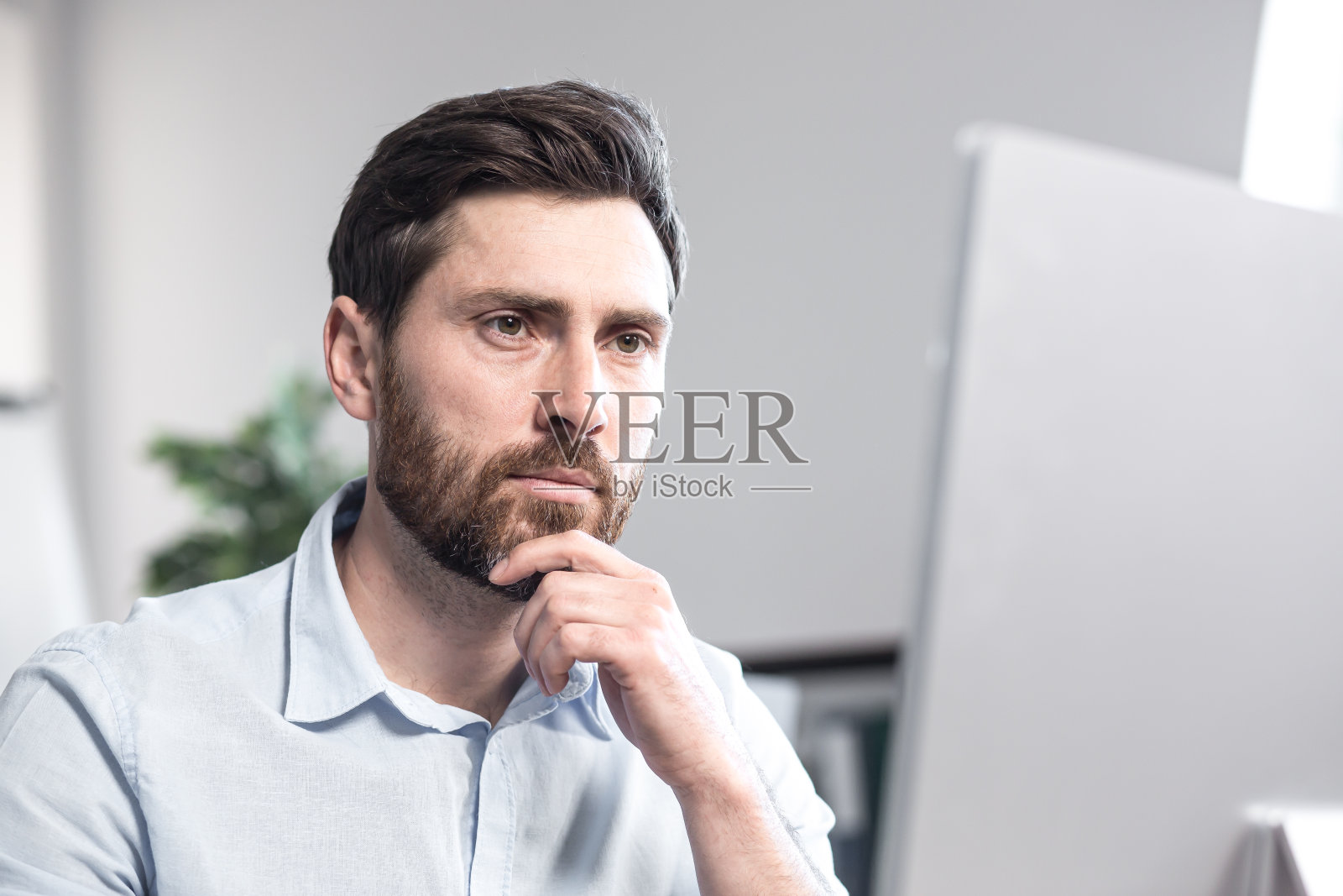 一个上班族的肖像。深思熟虑,一本正经,留着胡子。穿着白衬衫坐在办公室的电脑桌前。照片摄影图片