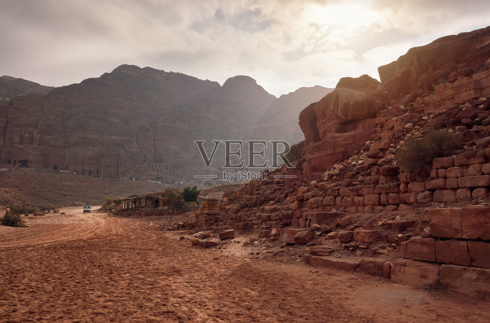 约旦佩特拉的典型景观:红色尘土飞扬的地面,远处是山脉和内部雕刻的建筑物照片摄影图片