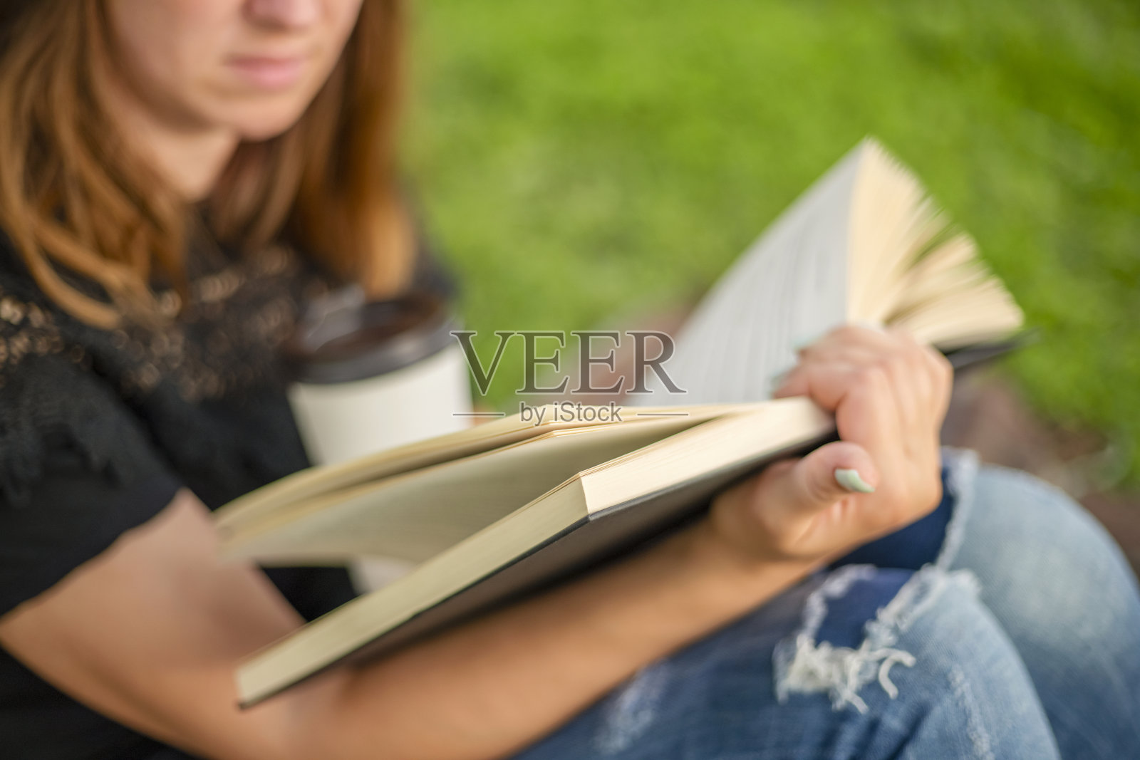 一个穿着牛仔裤的女人端着一杯咖啡坐在公园的毯子上看书照片摄影图片