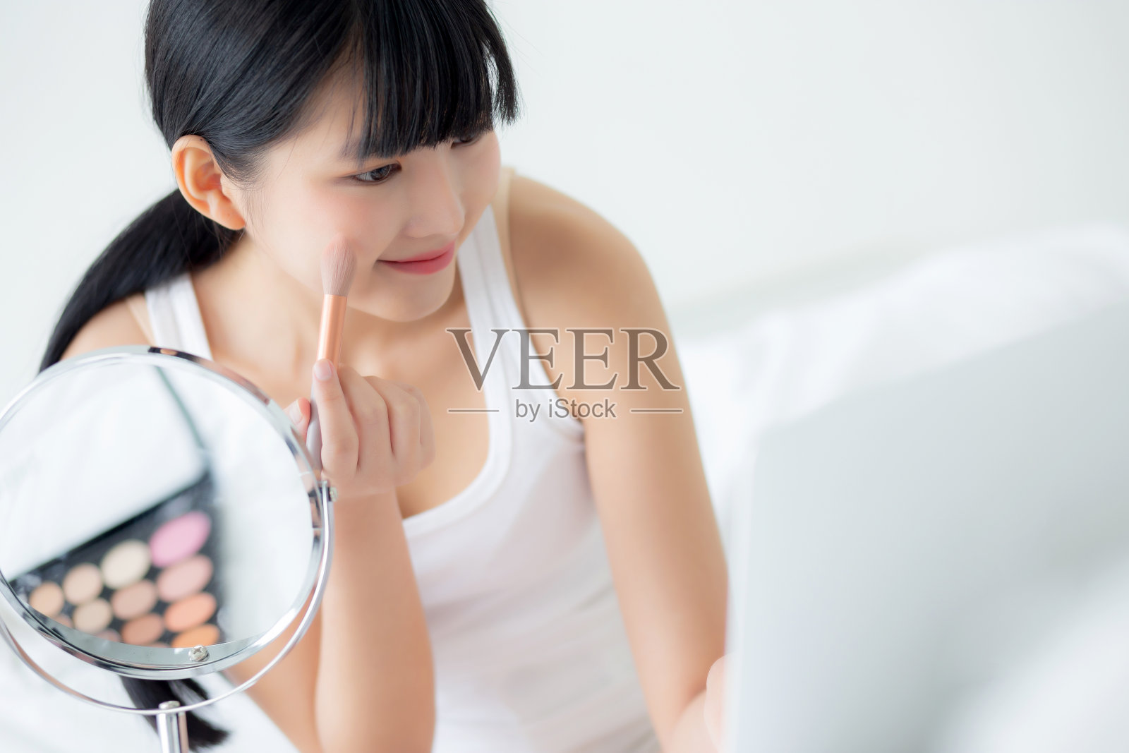 美丽的年轻亚洲女性与学习化妆刷脸颊在笔记本电脑上的教程在床上,女性教学和解释化妆与社交媒体流媒体。照片摄影图片