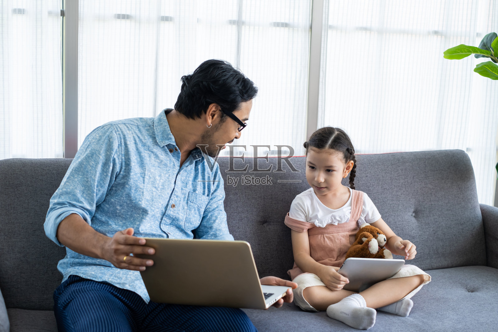 坐在客厅的沙发上,和爸爸一起玩,在家远程工作,用笔记本电脑在线学习,选择性地聚焦在女孩的脸上。家庭学校,父母。照片摄影图片
