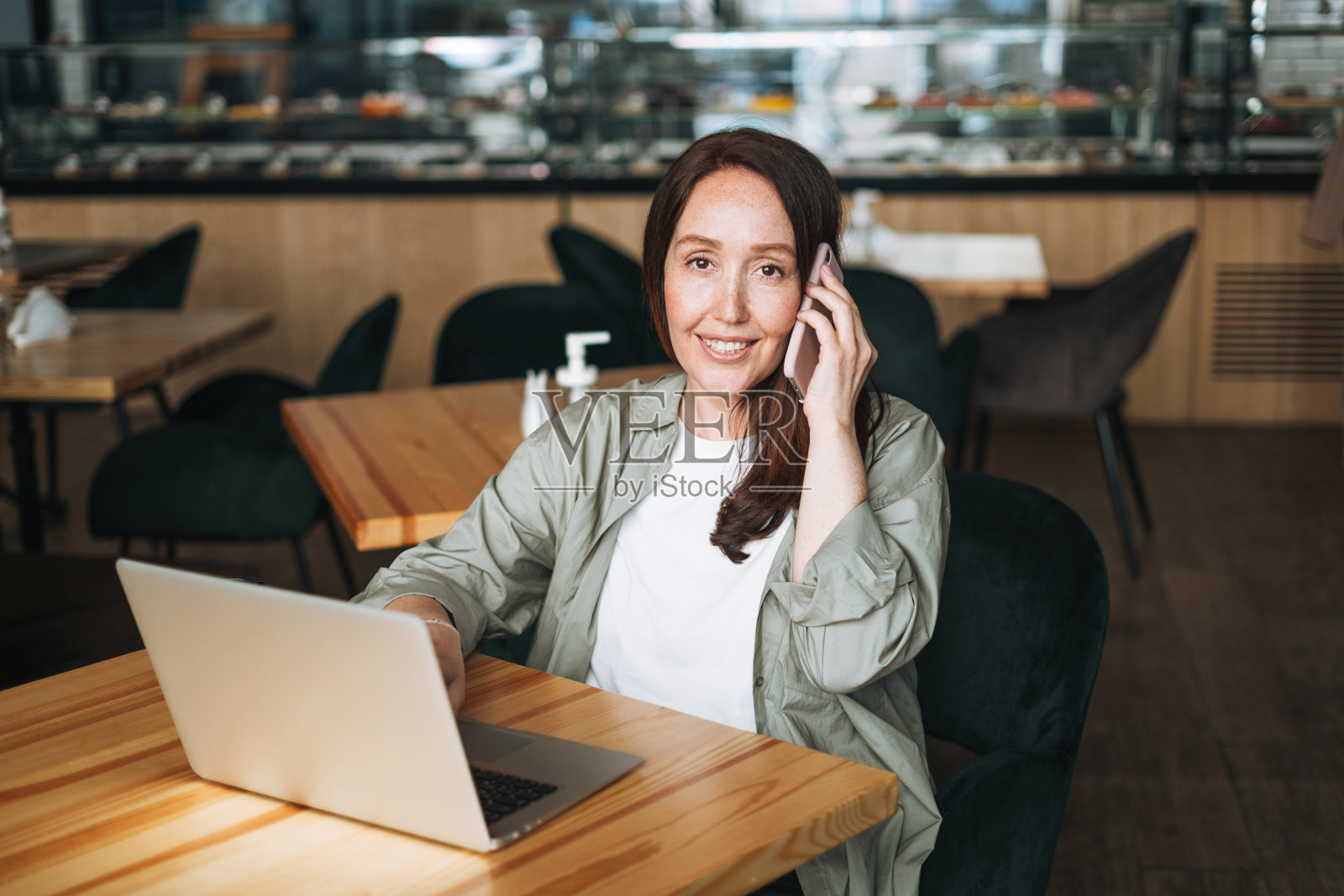 成人微笑的深褐色的商业女性40年的长发在时尚的衬衫在笔记本电脑使用手机在咖啡馆工作照片摄影图片