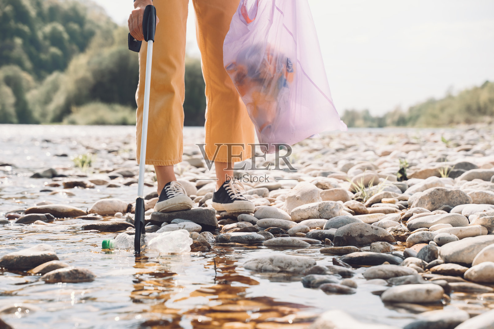 人们帮助保持大自然的清洁和捡垃圾。年轻女子清理室外的垃圾。女志愿者在河滩的石头上收集塑料垃圾。照片摄影图片