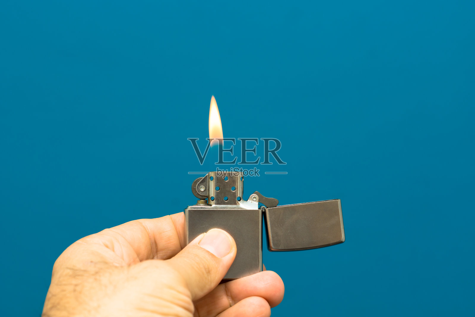 男子手持刷铬打火机在蓝色的背景。金属打火机点燃照片摄影图片