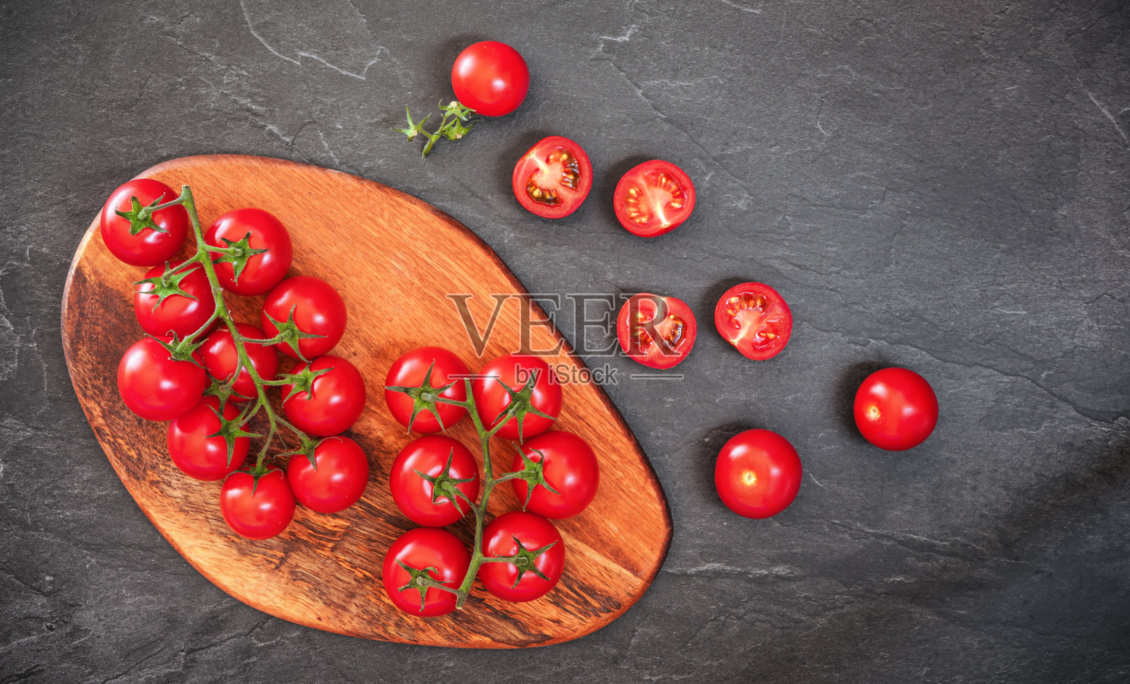 鲜红的圣女果放在木砧板上,有的切成两半放在黑色石板状砧板附近照片摄影图片