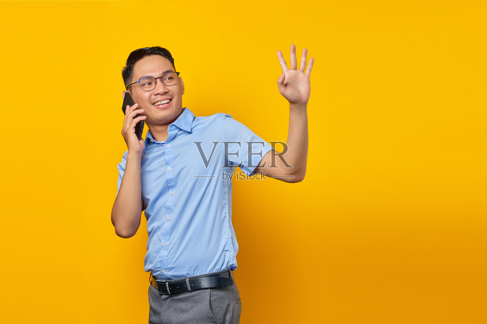 微笑的年轻亚洲人亚洲人在眼镜说话的手机,同时挥手手势孤立的黄色背景。商人与企业家概念照片摄影图片