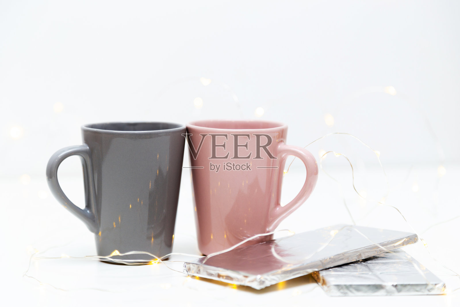 两个粉红色和灰色的咖啡杯模型和一块巧克力在白色的背景与小的金色灯光和bokeh。空白模板为您的设计,品牌,业务照片摄影图片