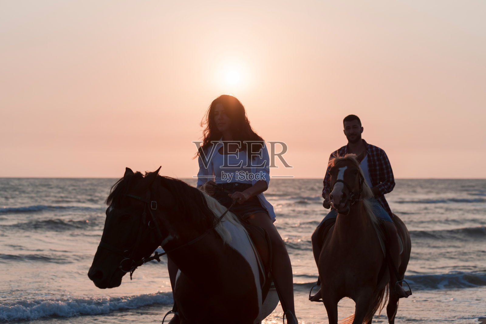 一对可爱的年轻夫妇穿着夏天的衣服,在夕阳下的沙滩上骑马。背景是大海和日落。有选择性的重点照片摄影图片