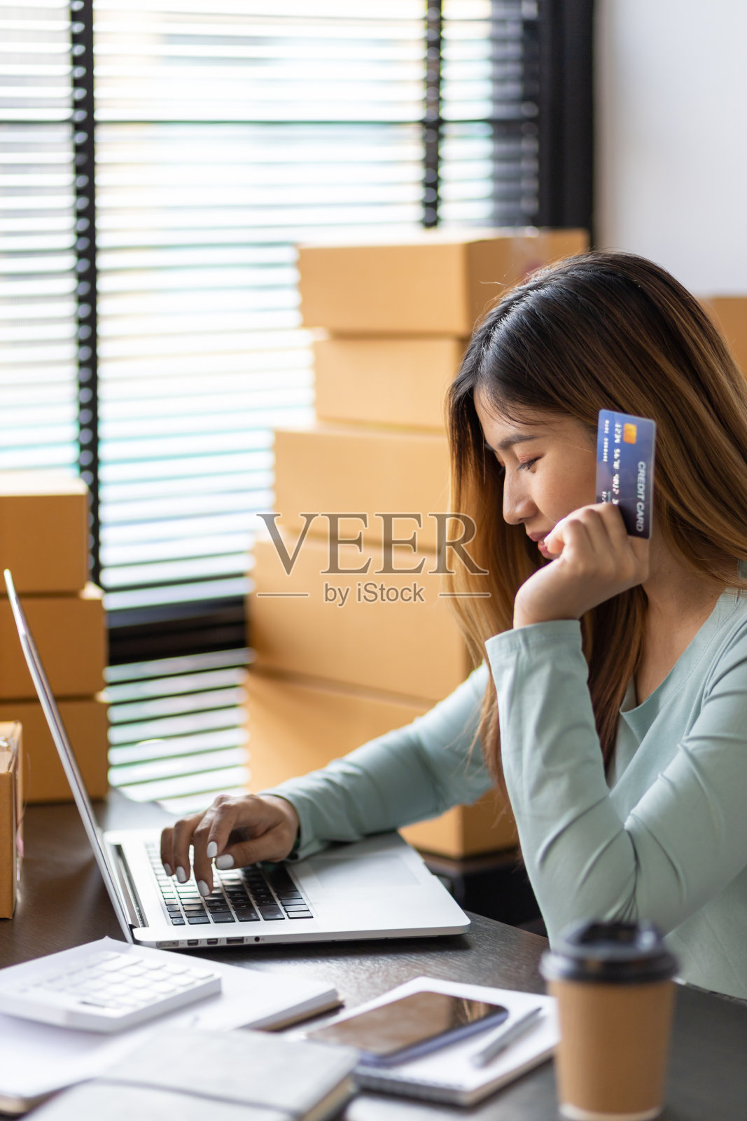 中小企业经营者正在用笔记本电脑检查网上订单的余额。小型企业的企业家持有信用卡,用笔记本电脑和家庭办公室的盒子工作。网上购物的女性。照片摄影图片